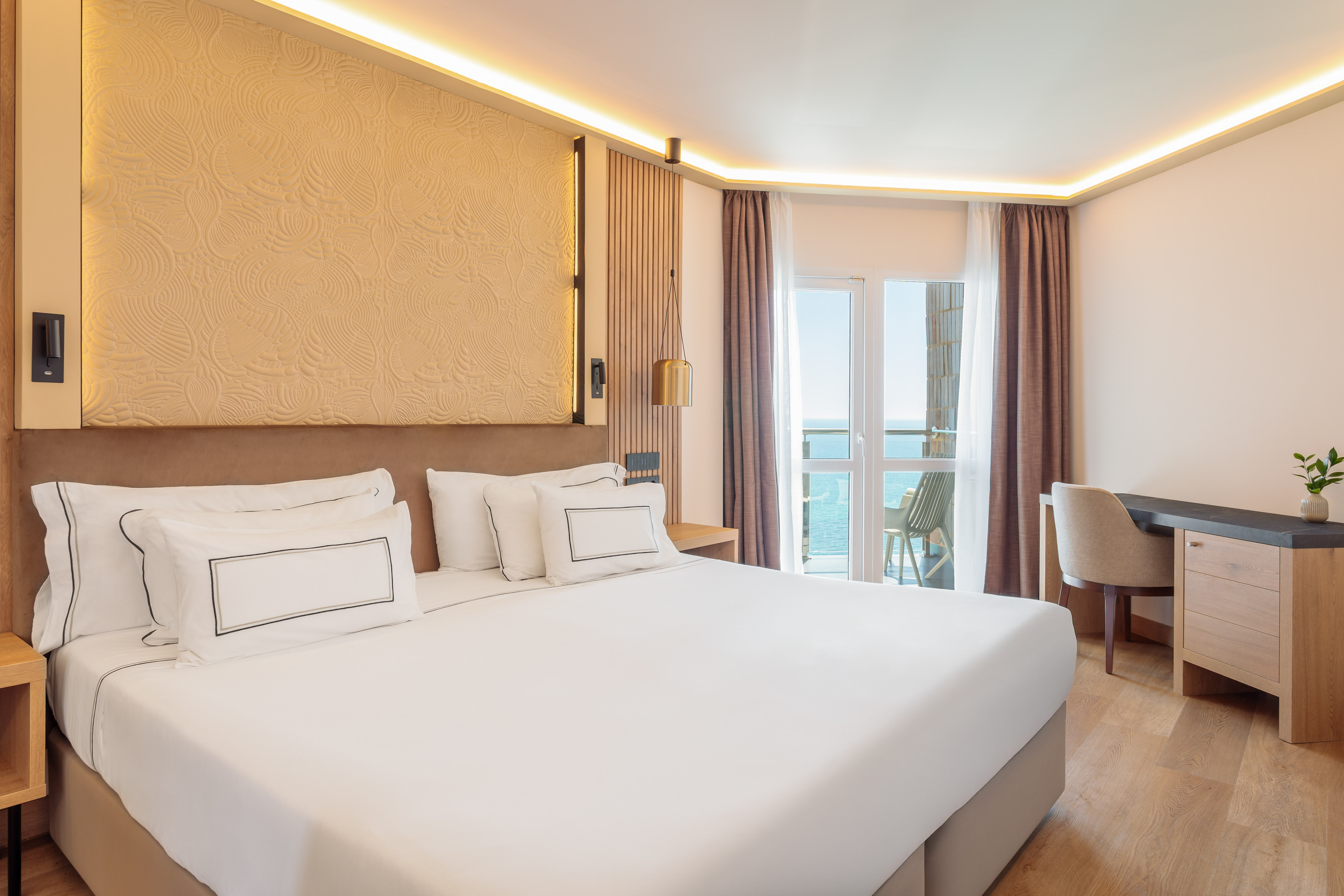 Modern hotel bedroom with king bed, sea view balcony, and wooden flooring.