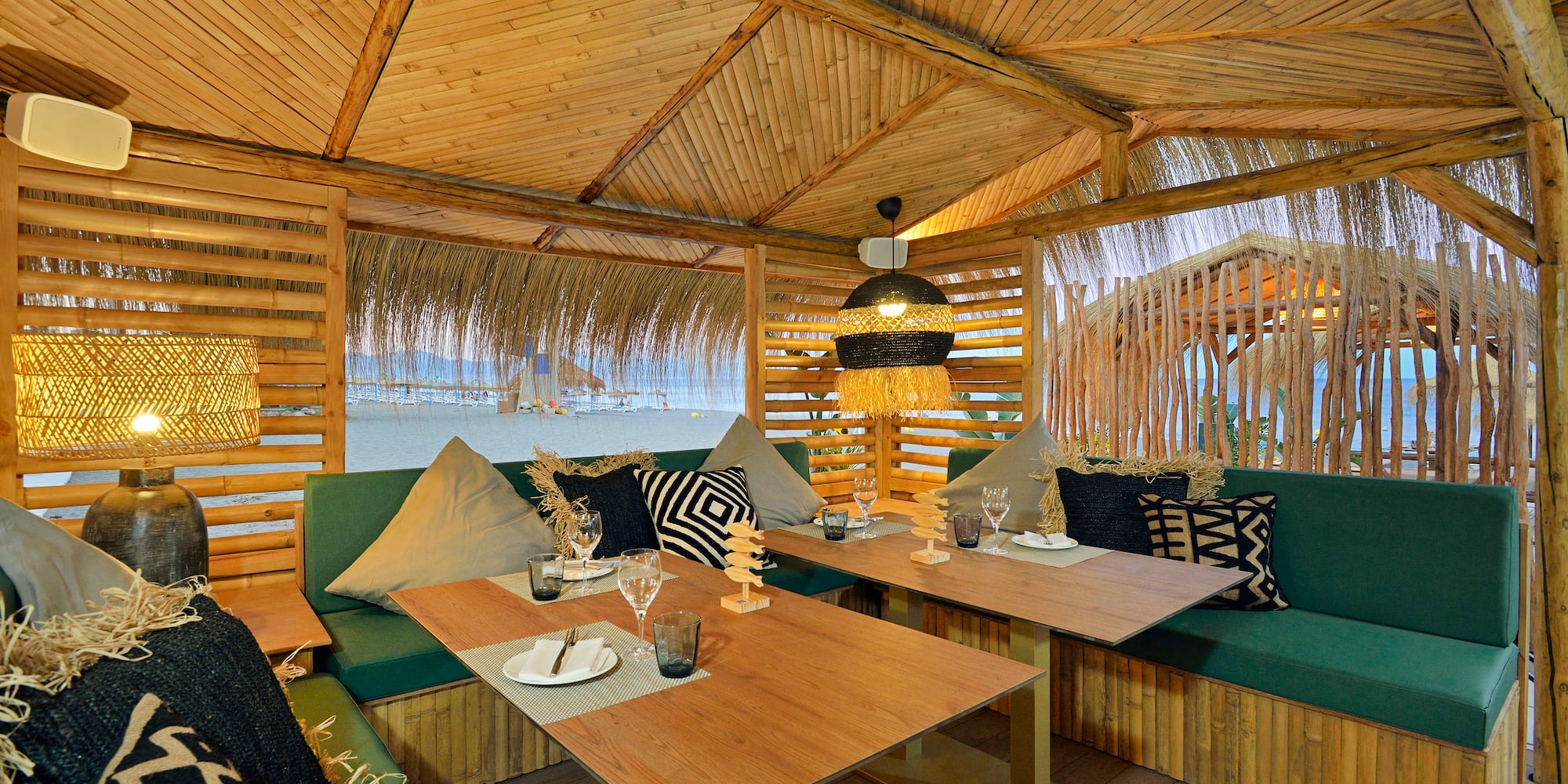 Bamboo beachside restaurant with green seating, wooden tables, and ocean view.