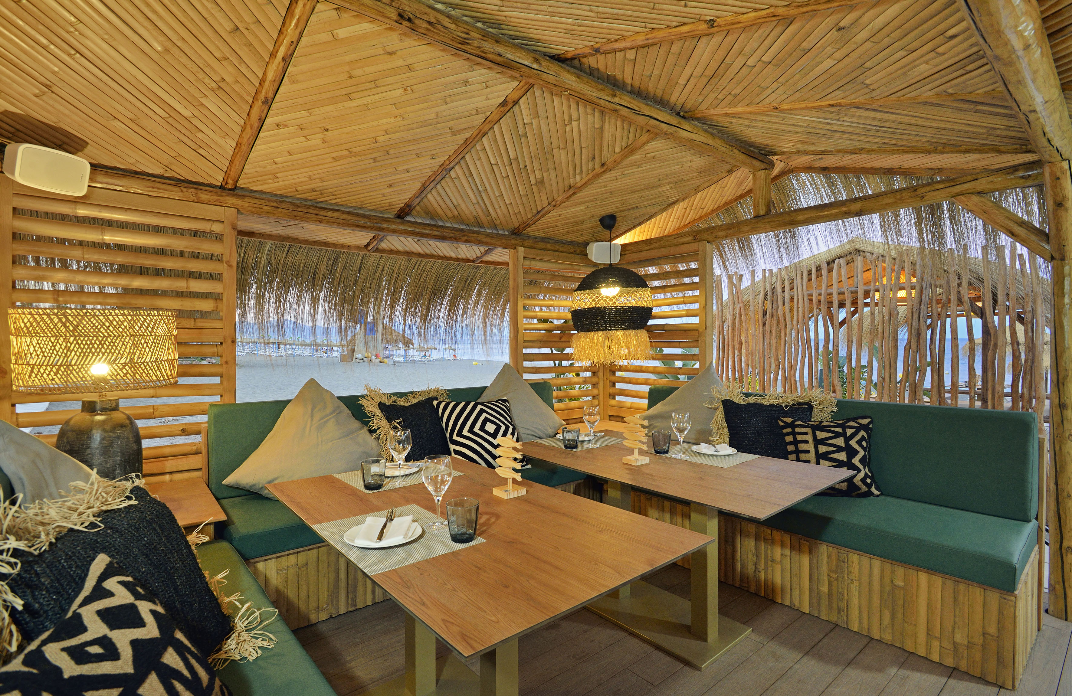 Bamboo beachside restaurant with green seating, wooden tables, and ocean view.