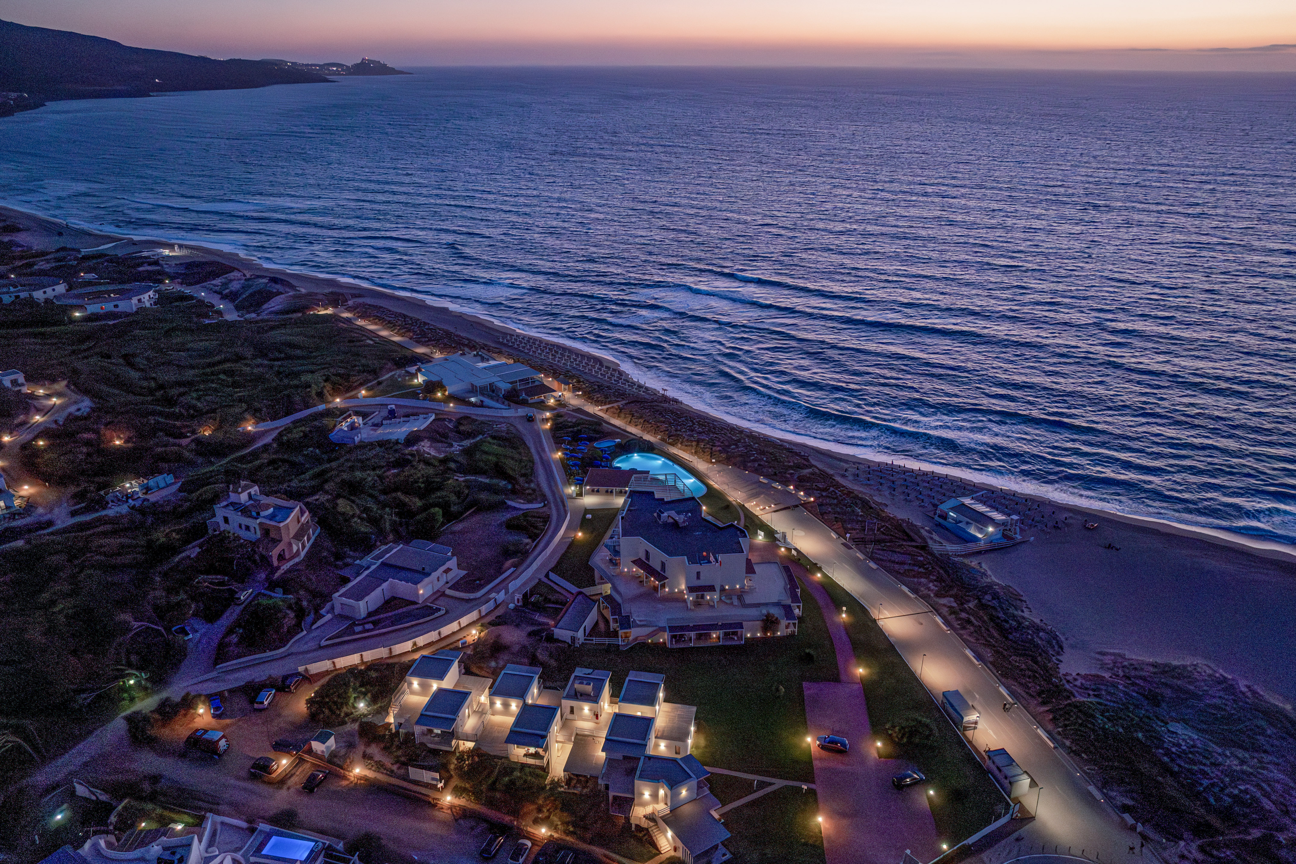 Aerial view of illuminated beach resort, ocean, and pool at twilight.