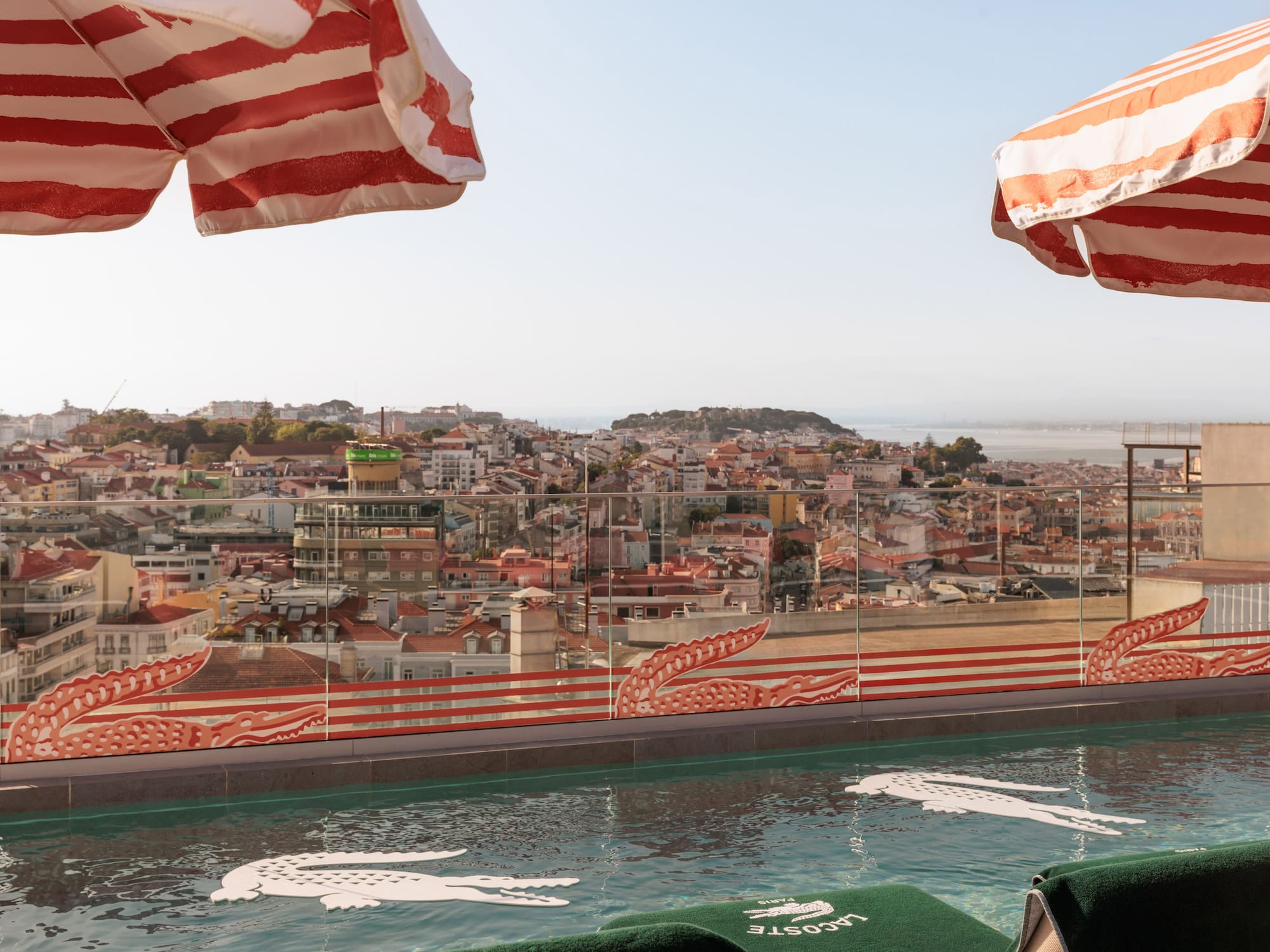 Lacoste rooftop pool with Lisbon skyline, crocodile floats, striped umbrellas.