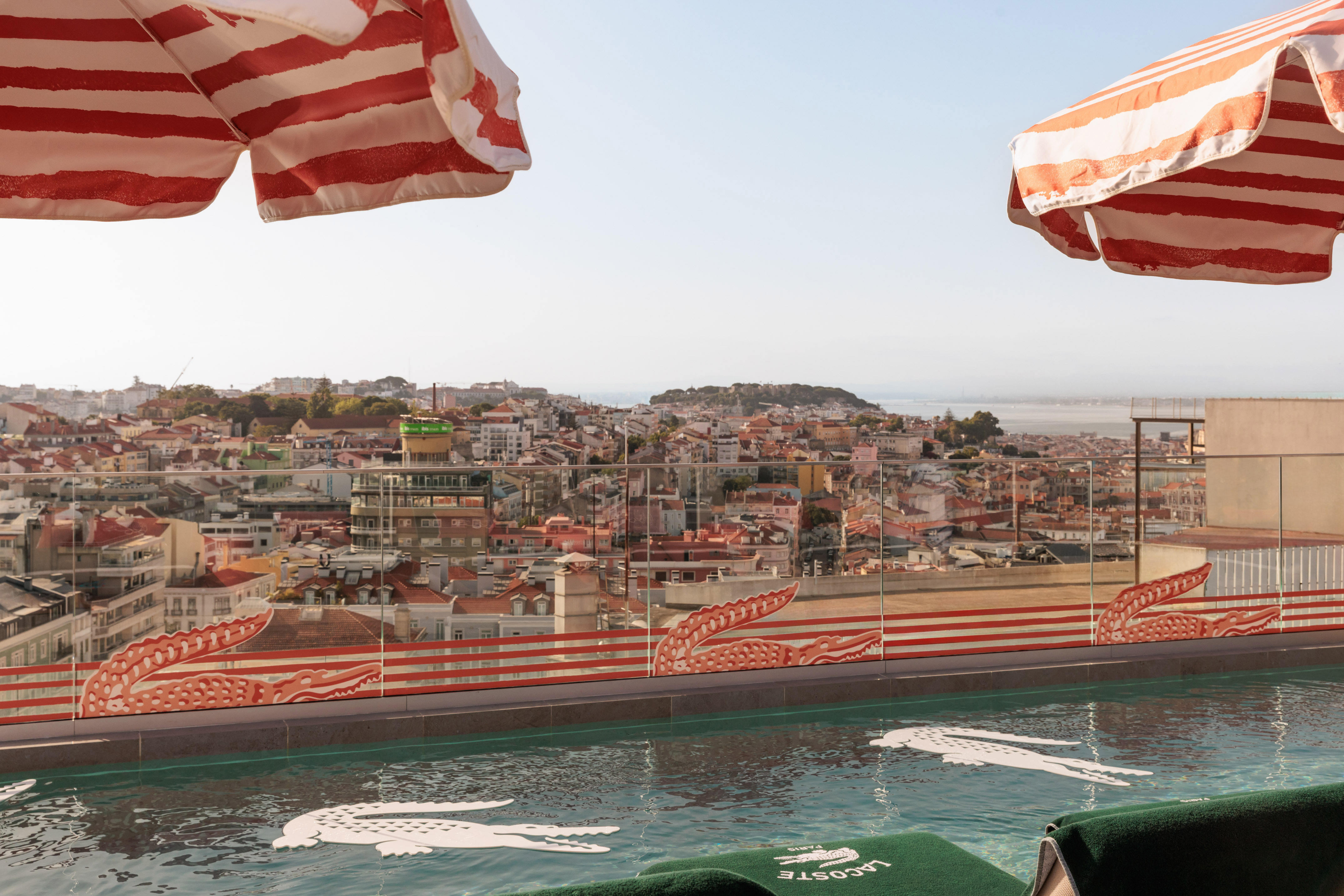 Lacoste rooftop pool with Lisbon skyline, crocodile floats, striped umbrellas.