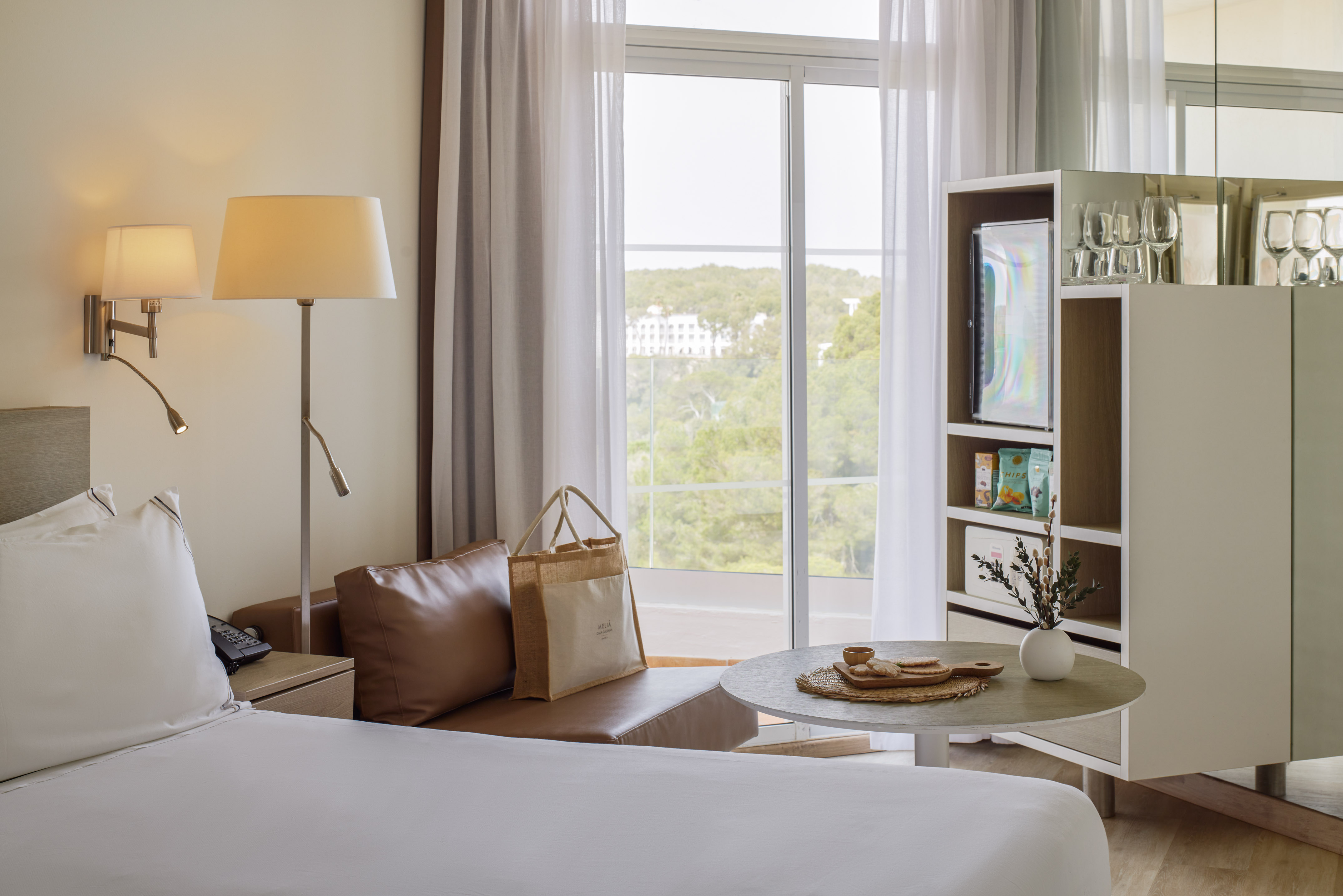 Modern hotel room with white bed, brown sofa, window, mini-bar, and resort view.