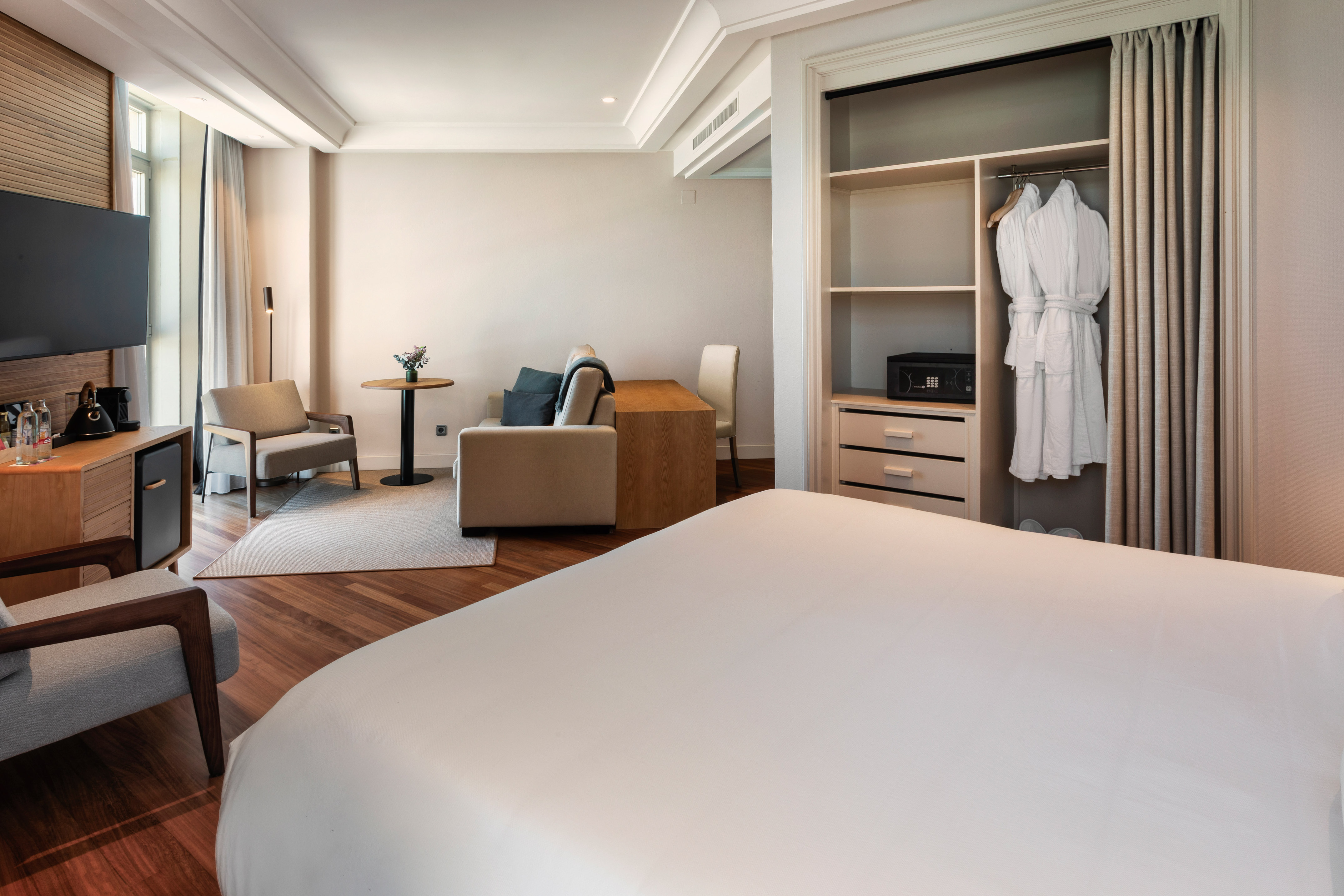 Hotel room with bed, sofa, TV, minibar, open closet, and bathrobes.