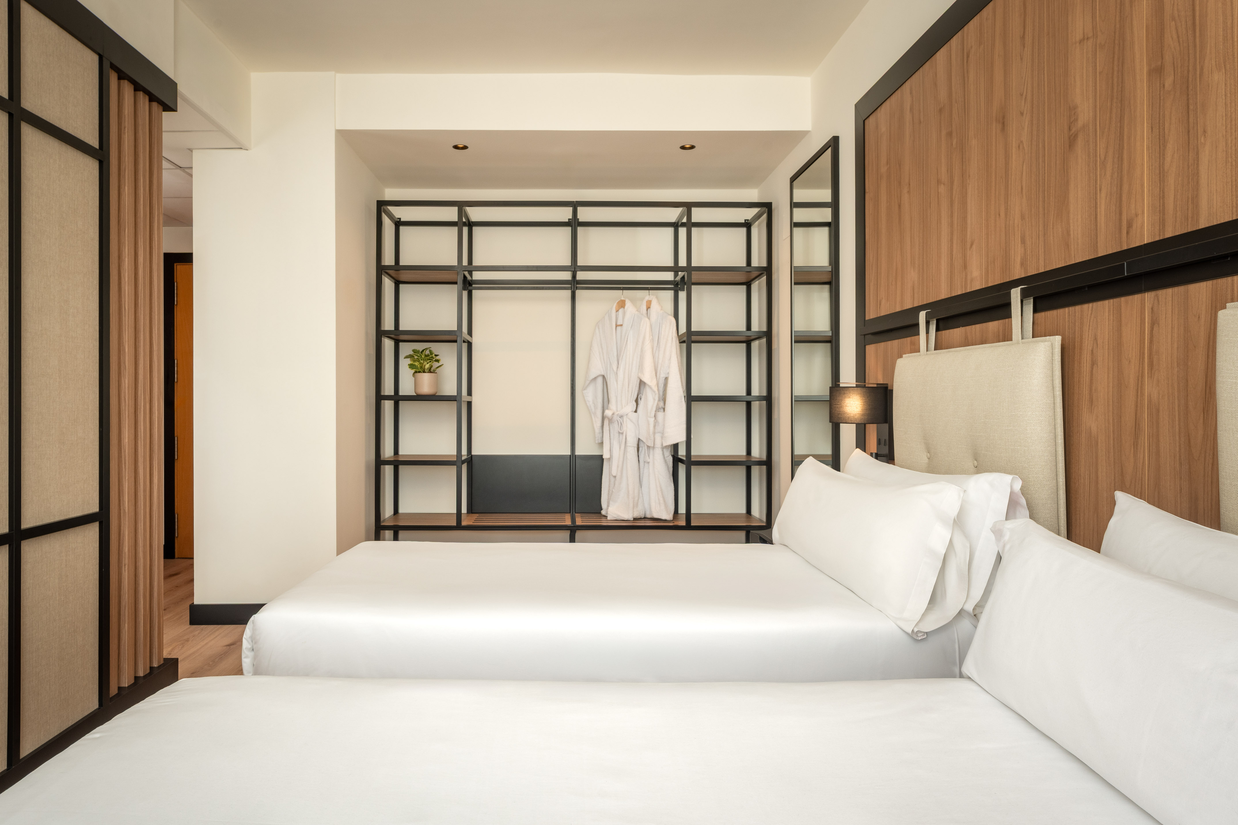 Modern hotel bedroom with two beds, open shelving unit, and white bathrobes.