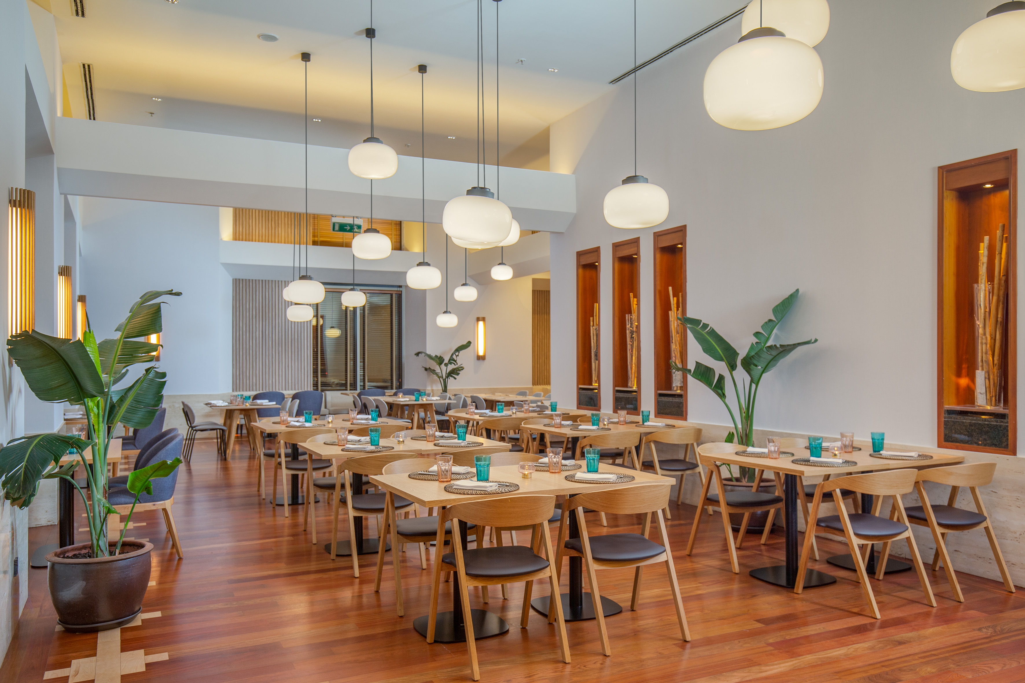 Modern restaurant with wooden dining tables, sleek pendant lights, and green plants.