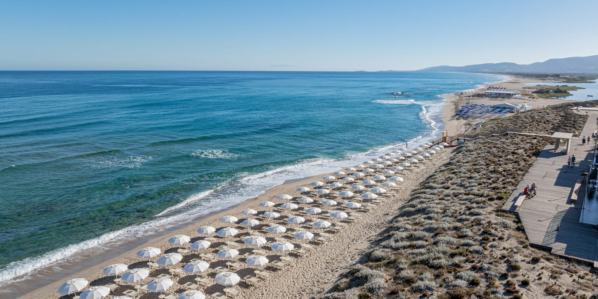 White umbrellas line a sandy beach with blue ocean and a coastal boardwalk.