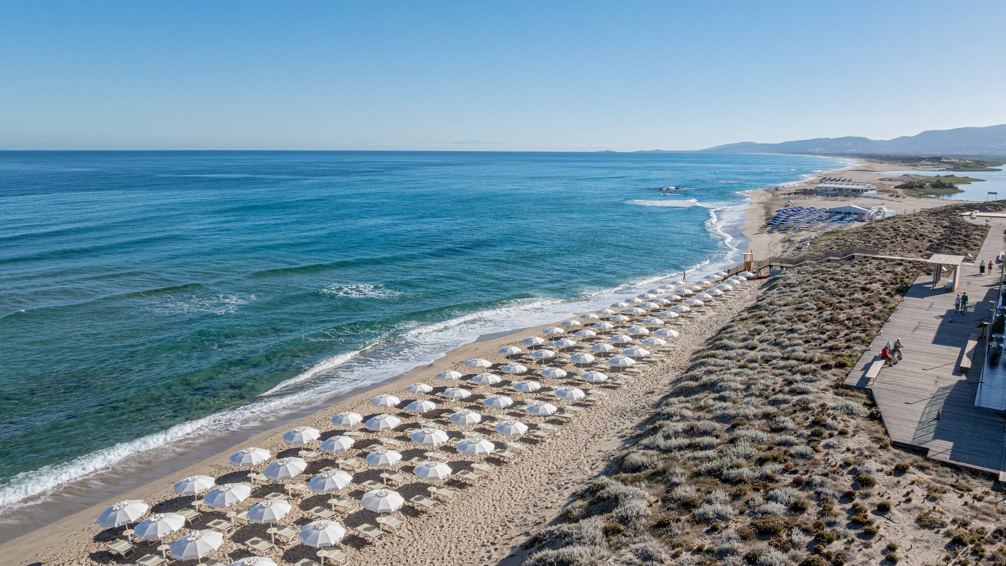 White umbrellas line a sandy beach with blue ocean and a coastal boardwalk.