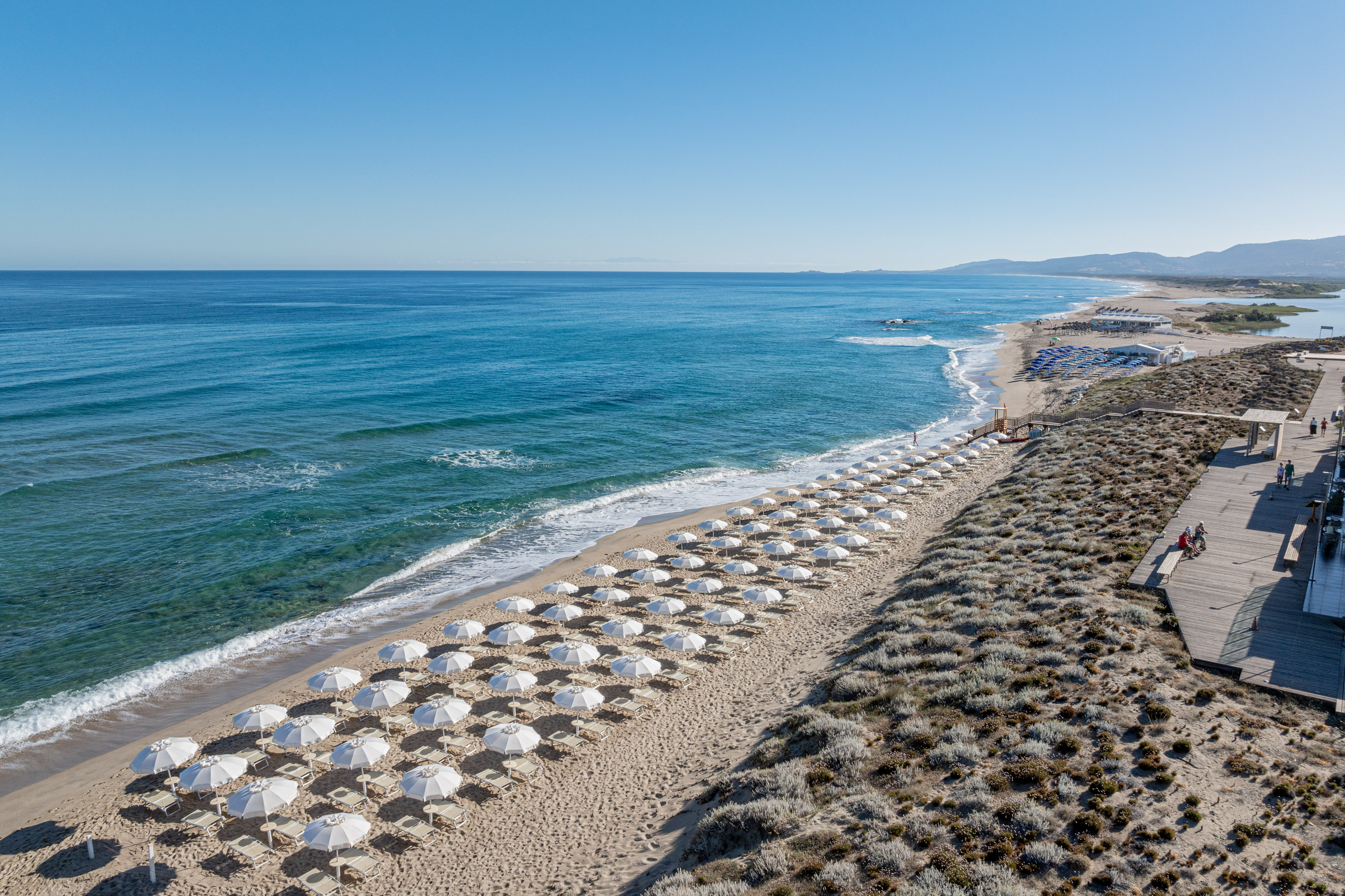 White umbrellas line a sandy beach with blue ocean and a coastal boardwalk.