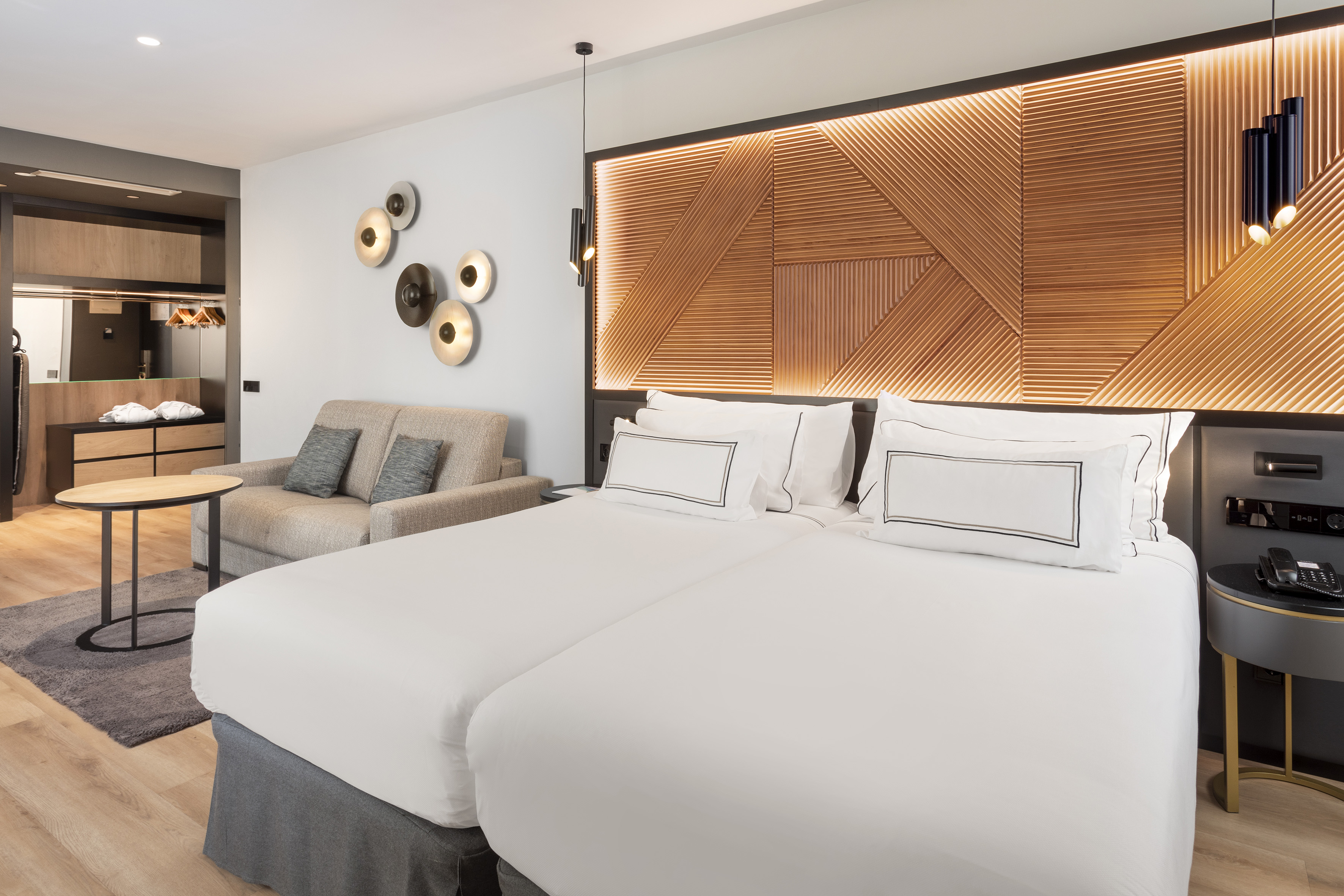 Modern hotel room with twin beds, sofa, and backlit wood panel headboard.