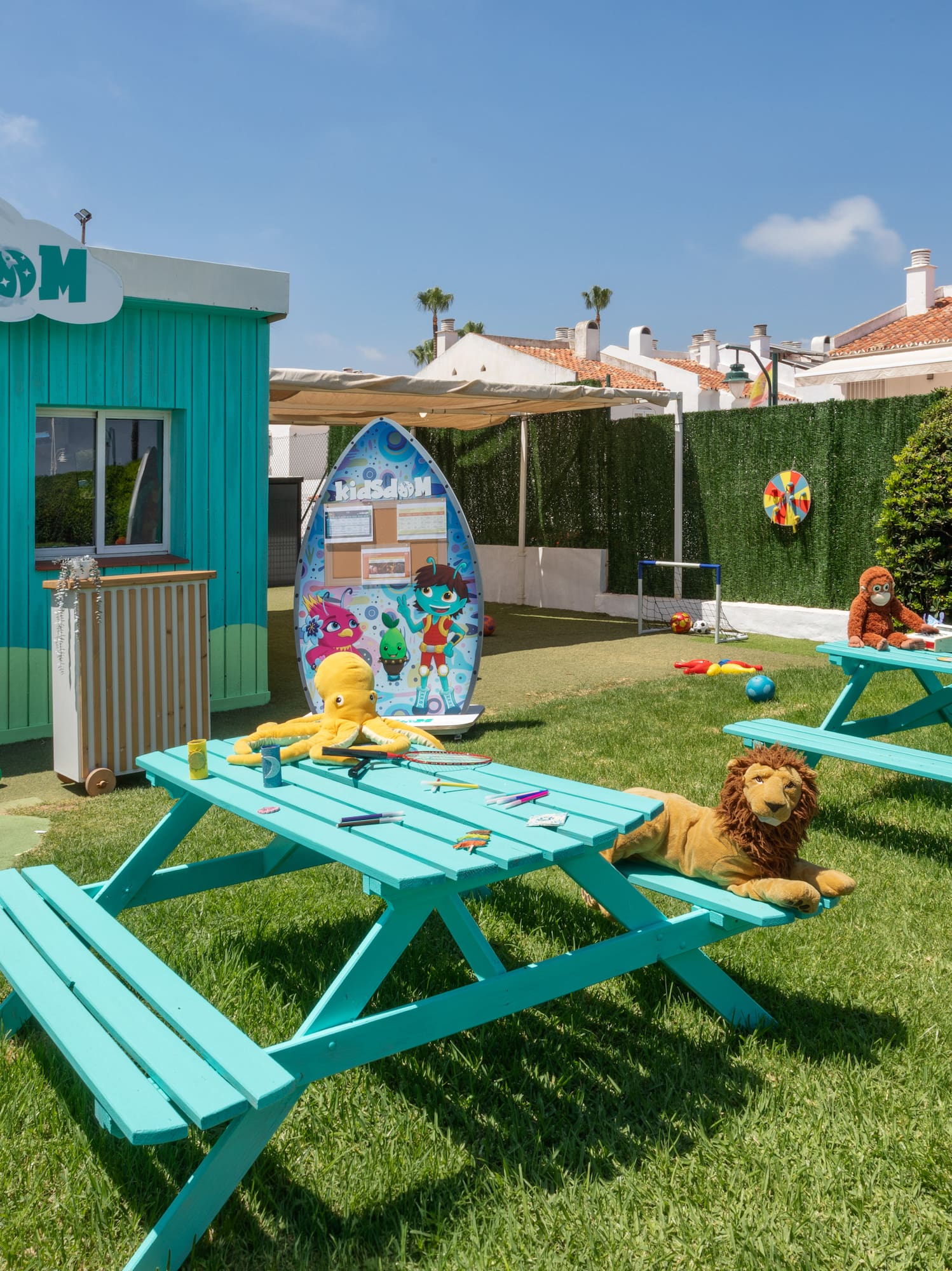 Área de juegos KidsDom exterior con mesas turquesa, peluches y césped.