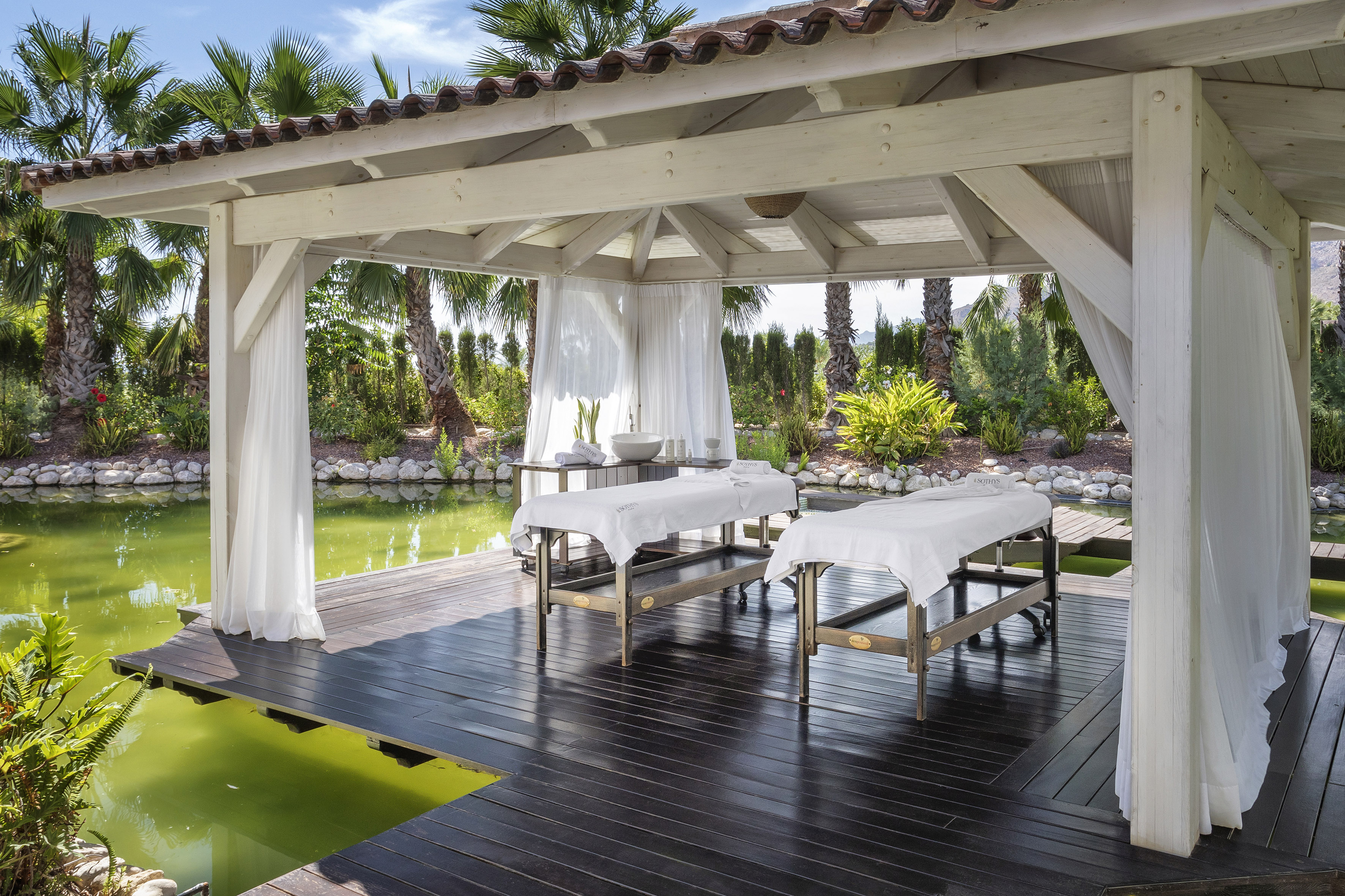 Outdoor spa gazebo with two massage tables by a pond and palm trees.