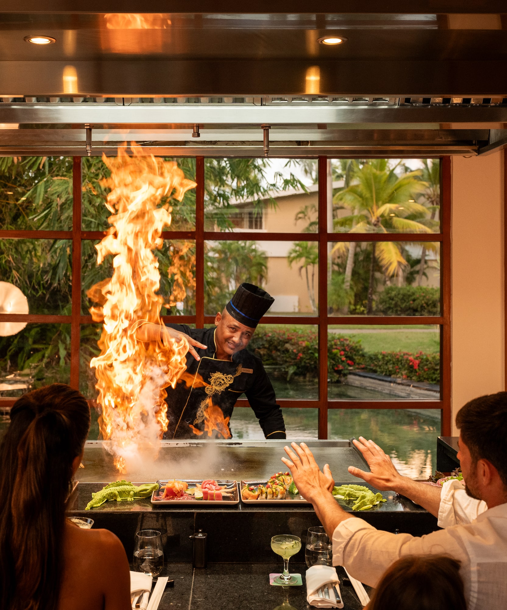 a man in a black hat cooking on a grill with a fire in front of a group of people