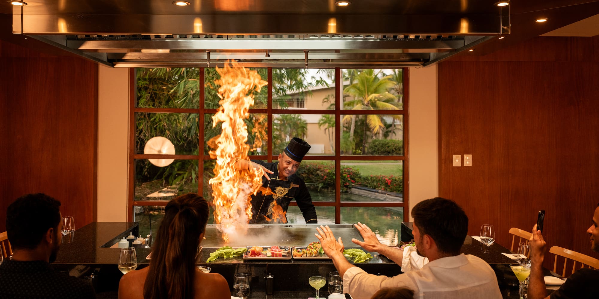 a man in a black hat cooking on a grill with a fire in front of a group of people