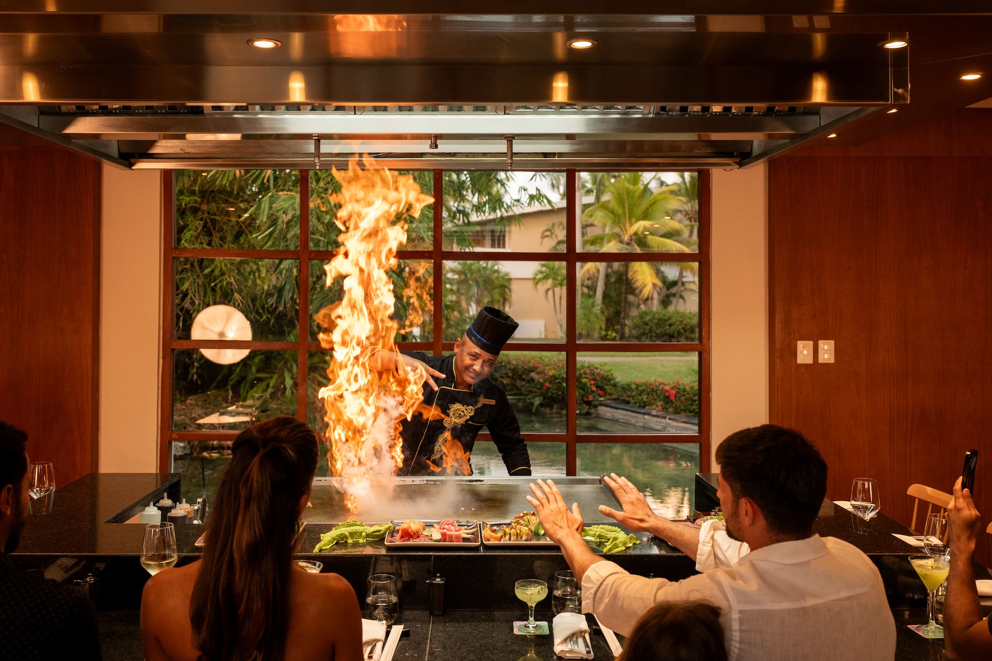 a man in a black hat cooking on a grill with a fire in front of a group of people