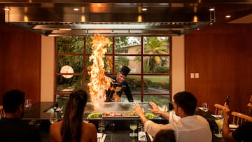 a man in a black hat cooking on a grill with a fire in front of a group of people