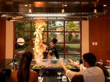 a man in a black hat cooking on a grill with a fire in front of a group of people