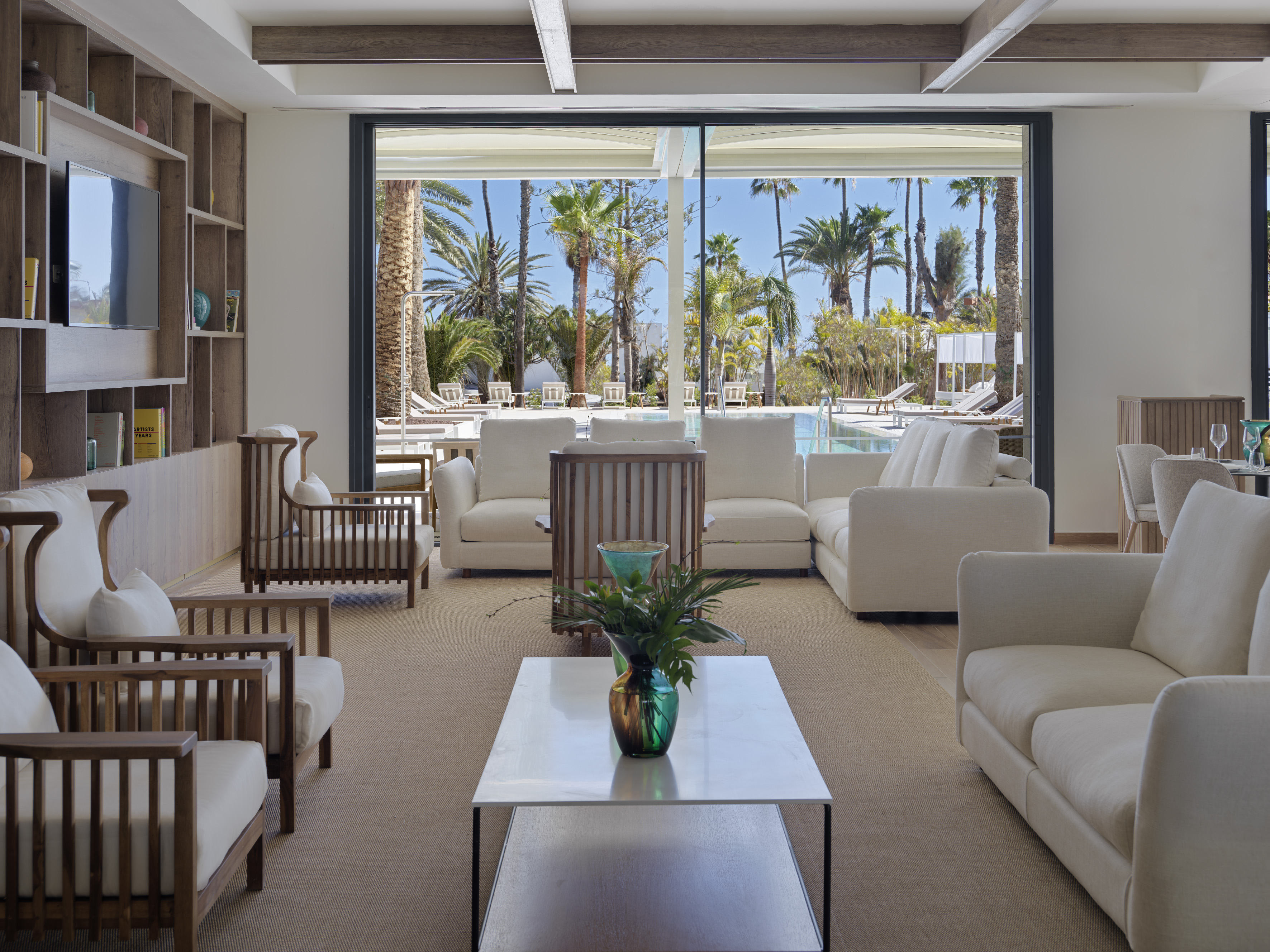 Hotel lounge with pool view, palm trees, white sofas, and wooden accents.