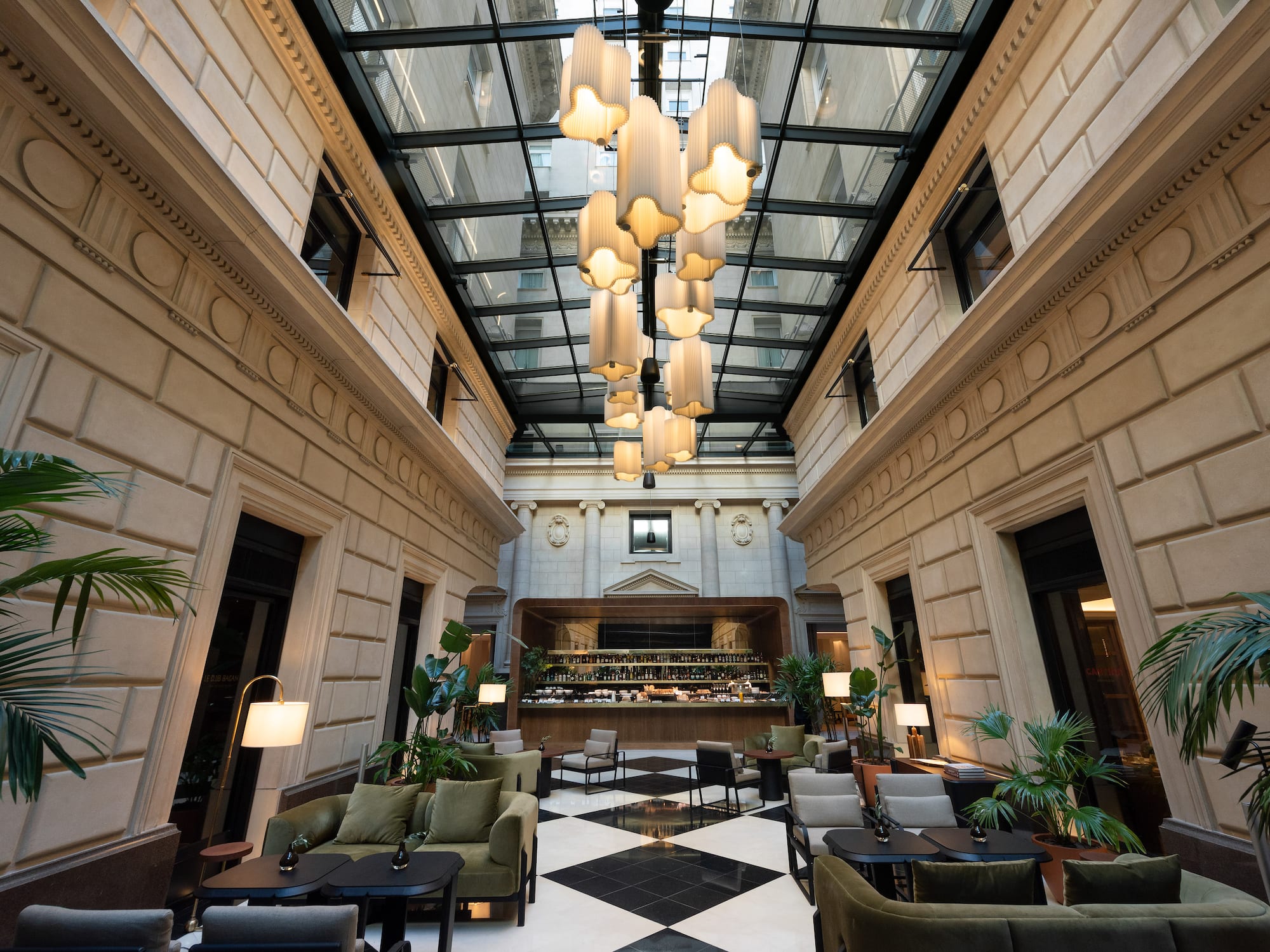 Hotel atrium lounge: glass roof, modern chandeliers, bar, and checkerboard floor.