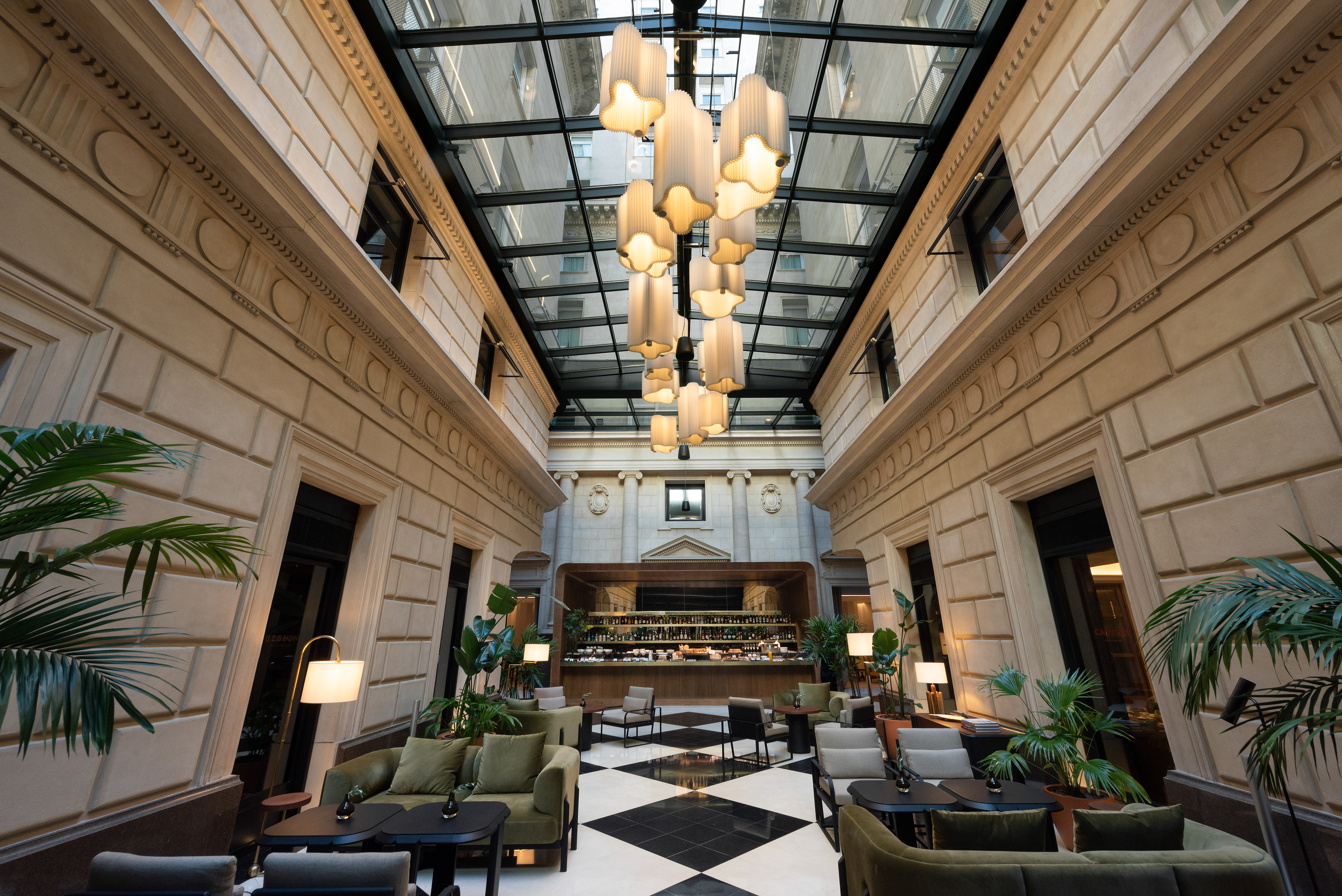 Hotel atrium lounge: glass roof, modern chandeliers, bar, and checkerboard floor.
