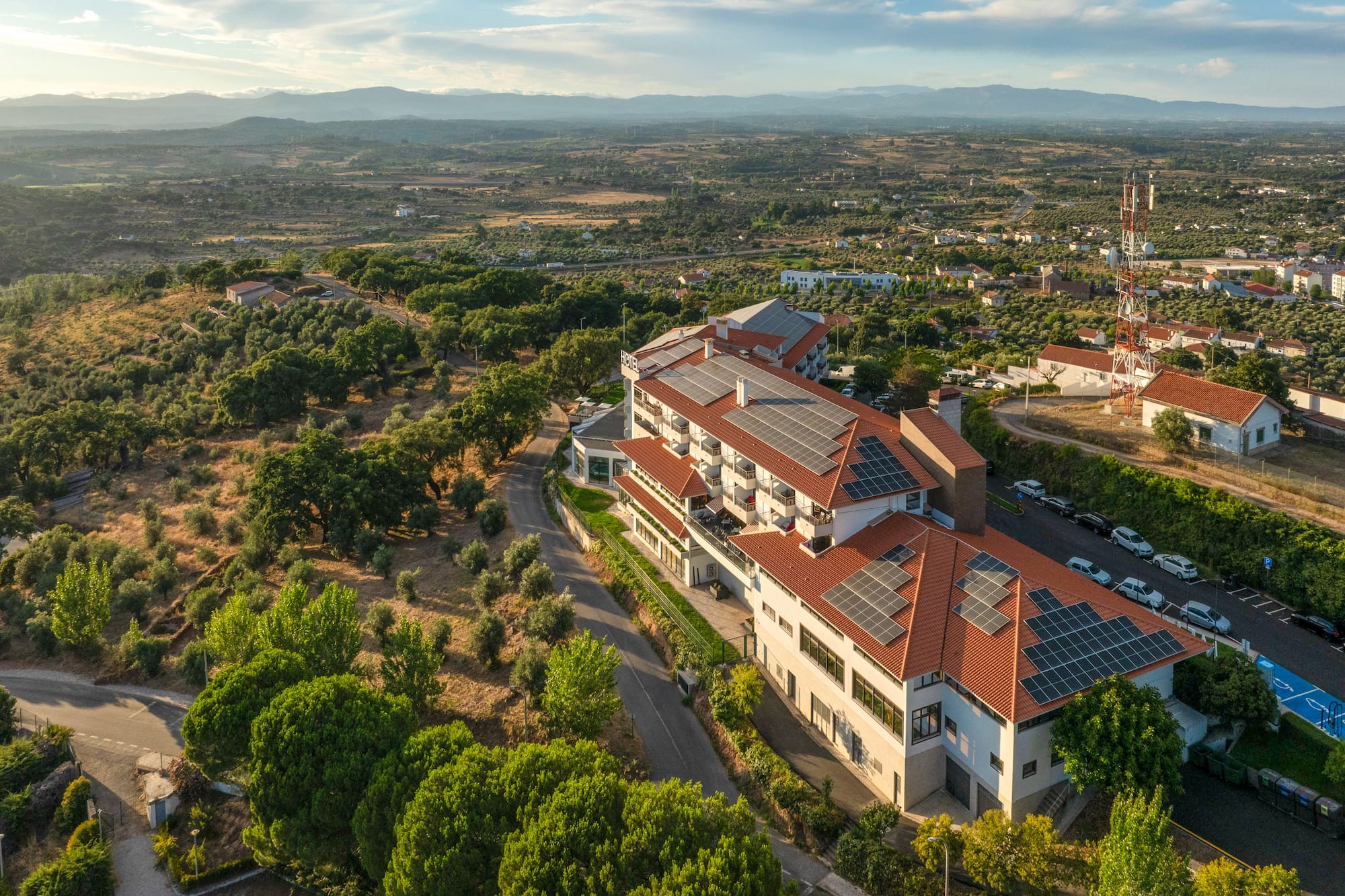 Aerial view of hotel with solar panels on red roofs, surrounded by green landscape.