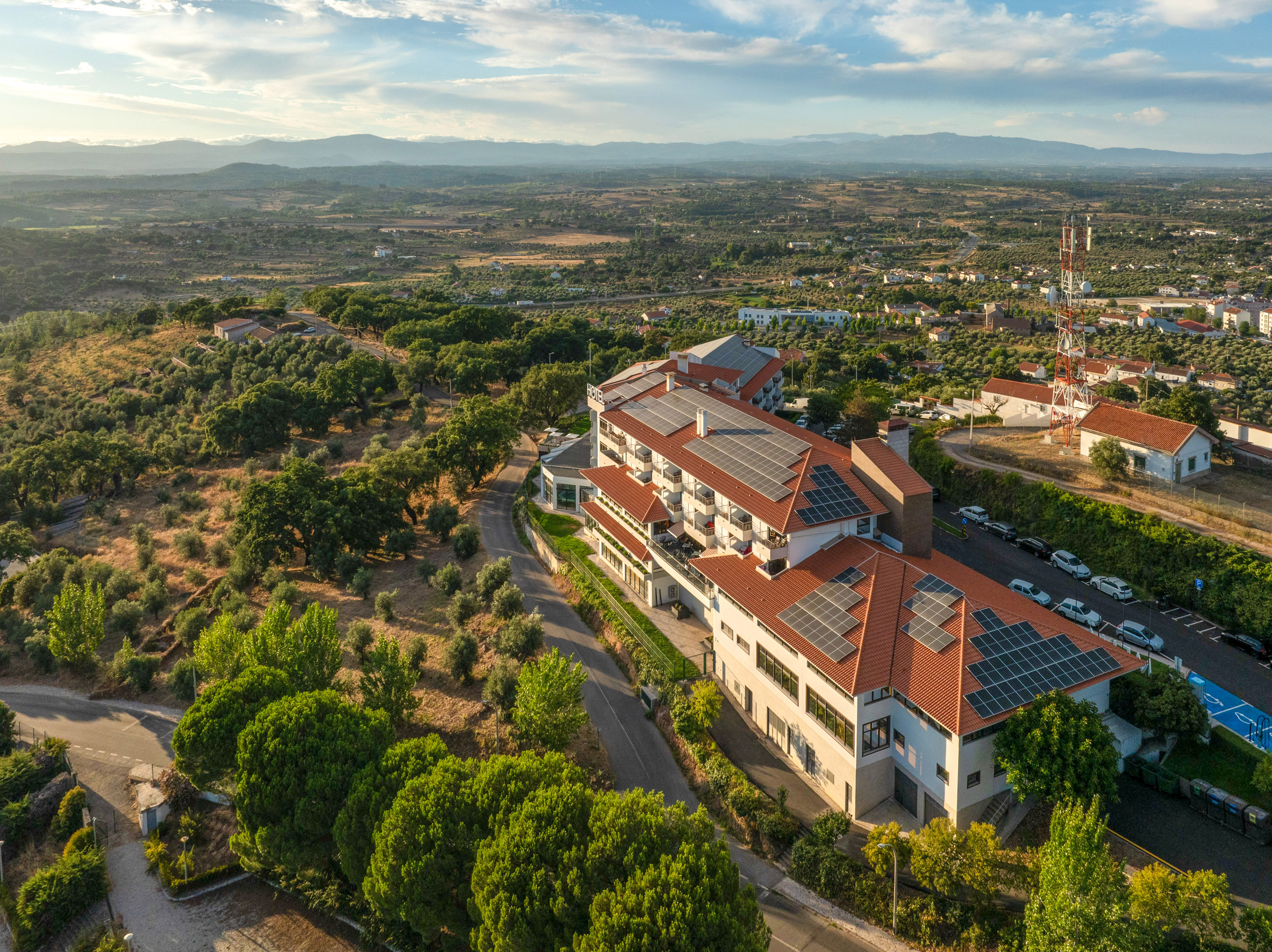 Aerial view of hotel with solar panels on red roofs, surrounded by green landscape.