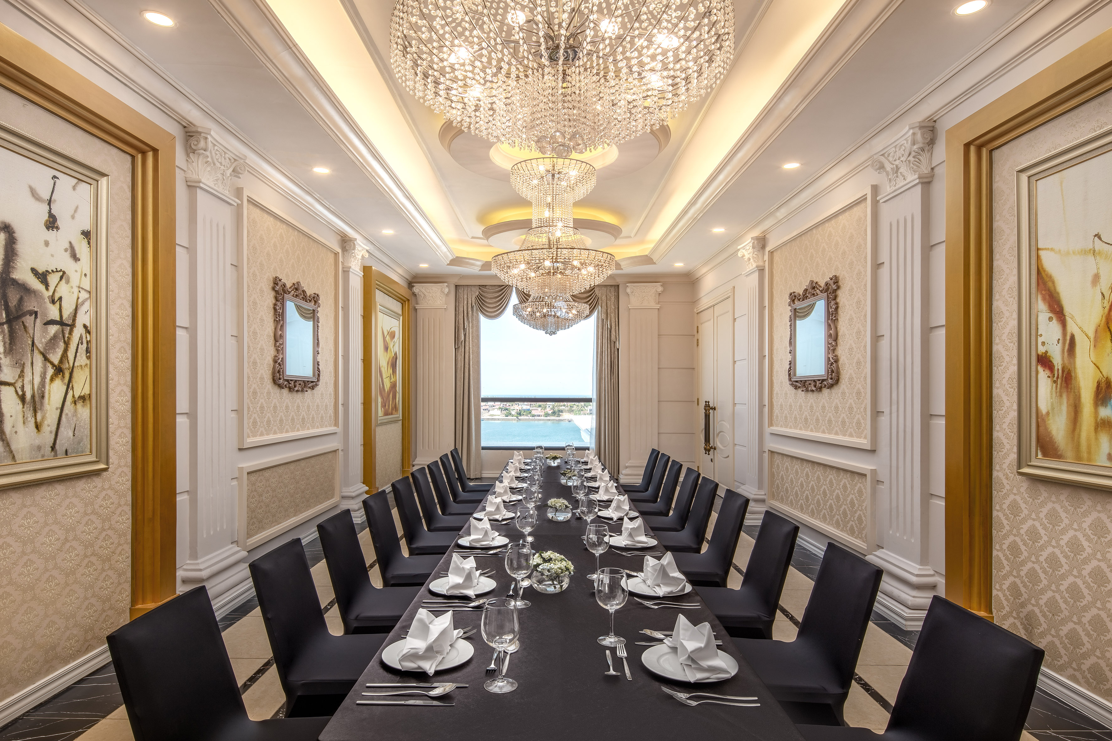 Luxury dining room with long formal table, crystal chandeliers, and water view.