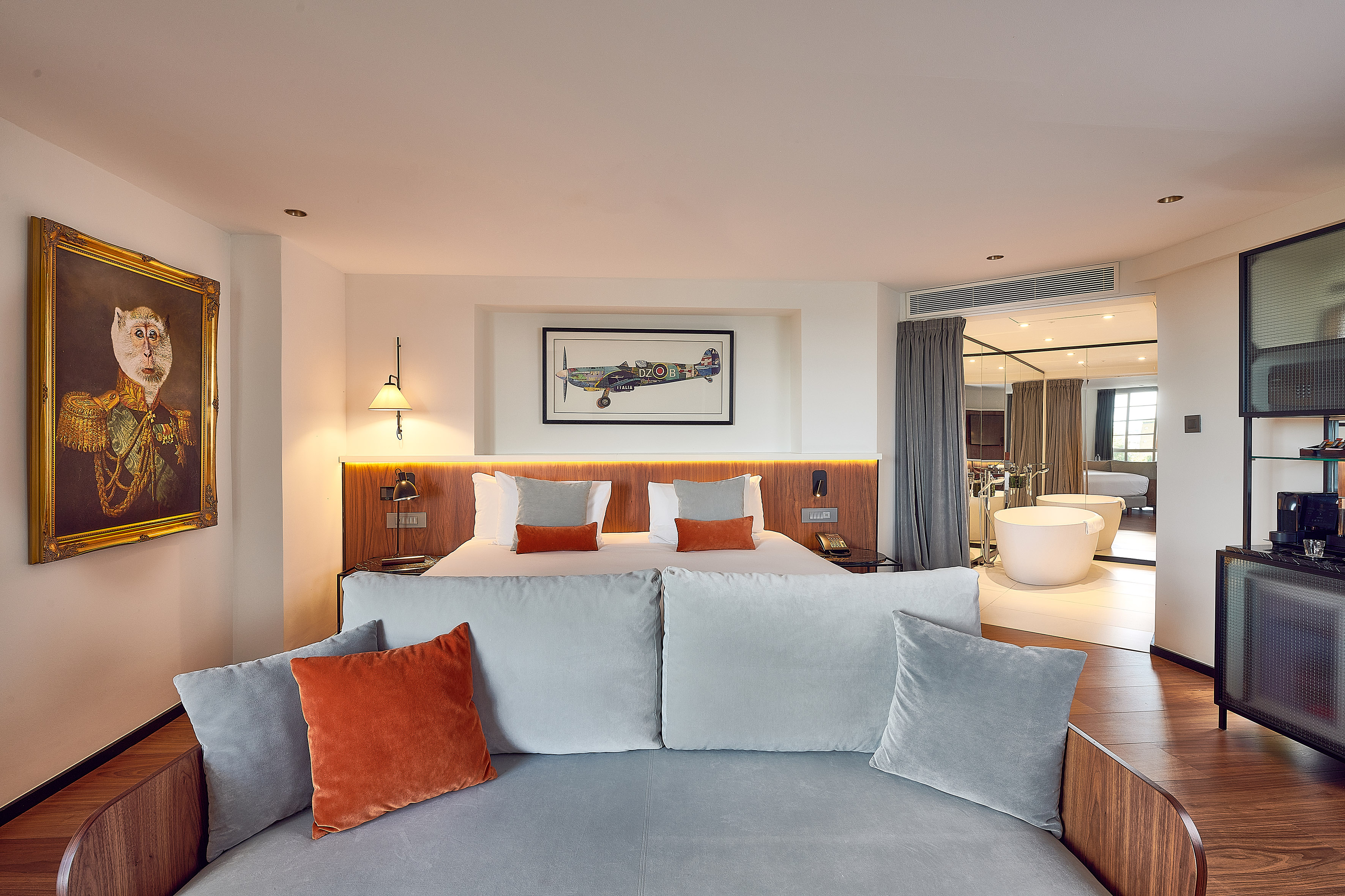 Luxury hotel suite with king bed, grey sofa, open-plan bathroom, and unique art.
