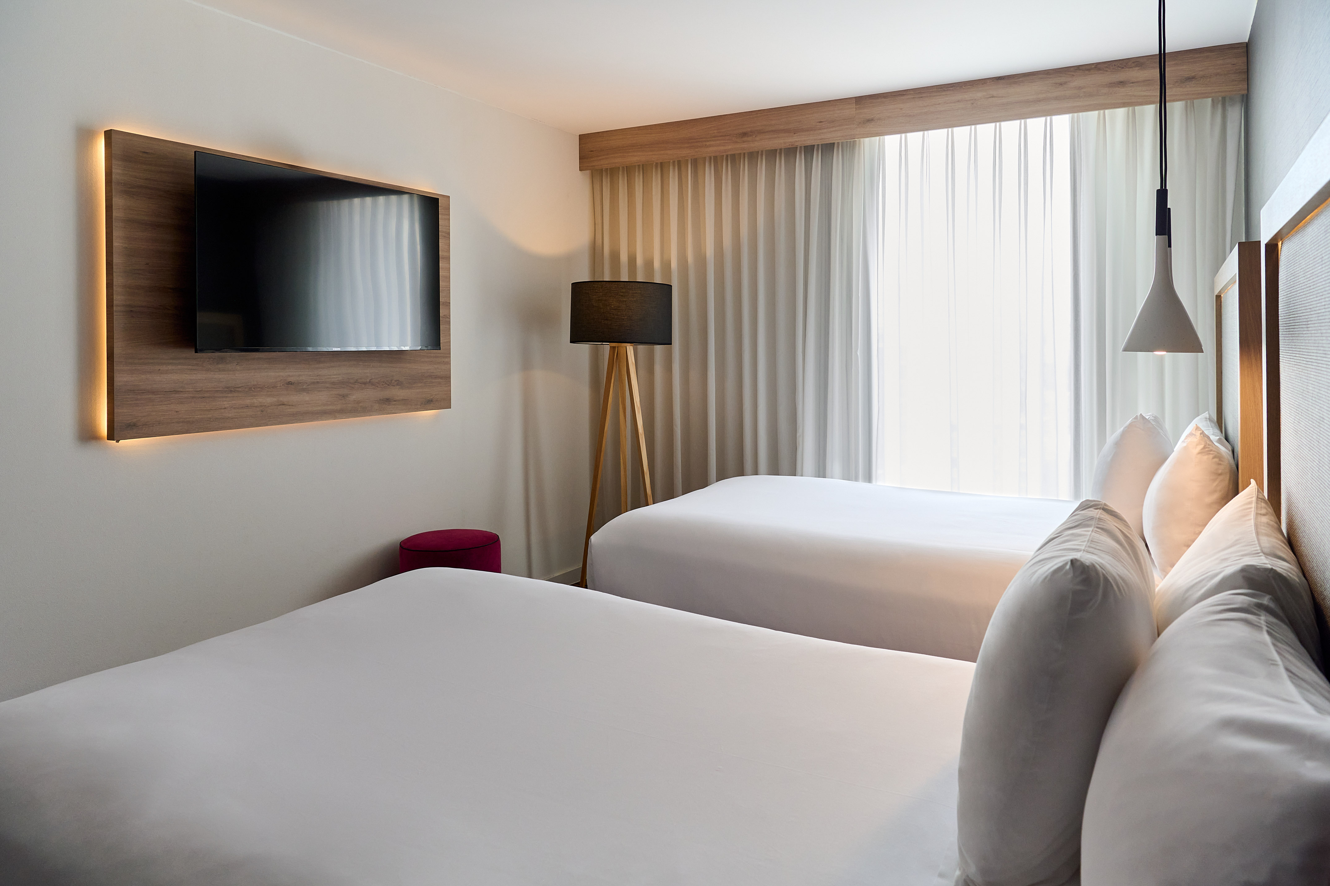Modern hotel room with back-lit TV, twin beds, tripod lamp, and sheer curtains.