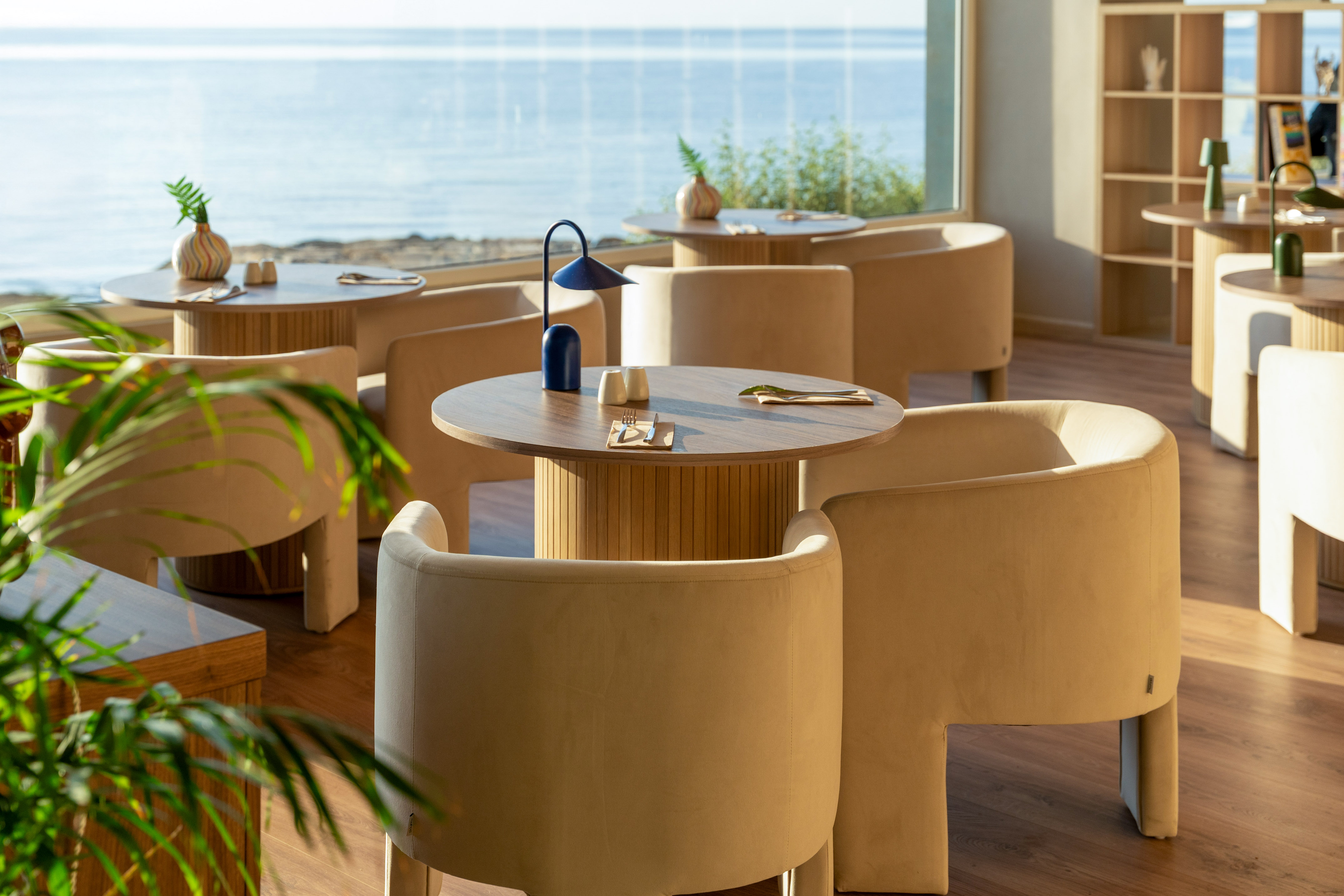 Coastal restaurant interior with sea view, wooden tables, and beige chairs.