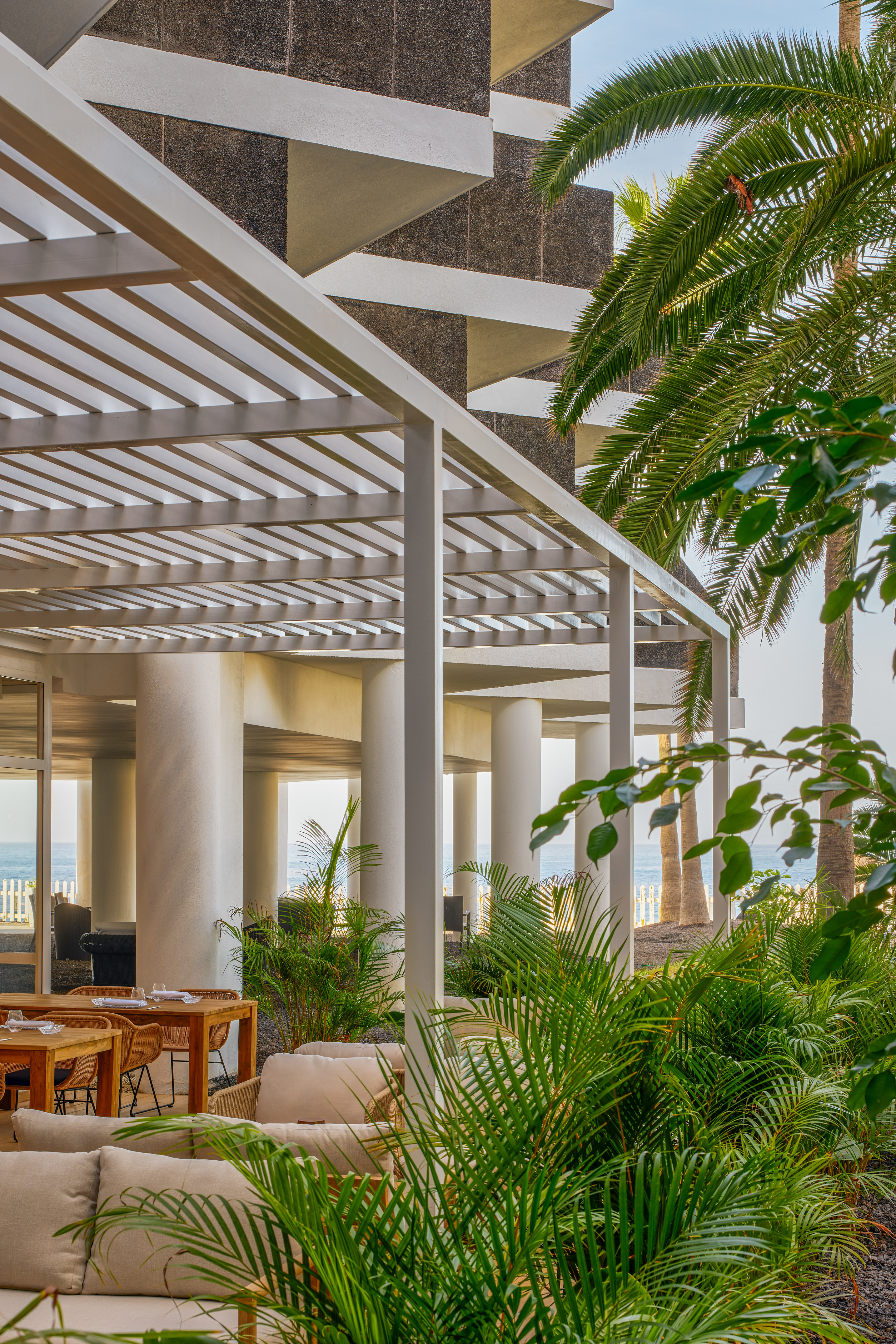 Tropical outdoor restaurant patio with white pergola, palm trees, ocean view.