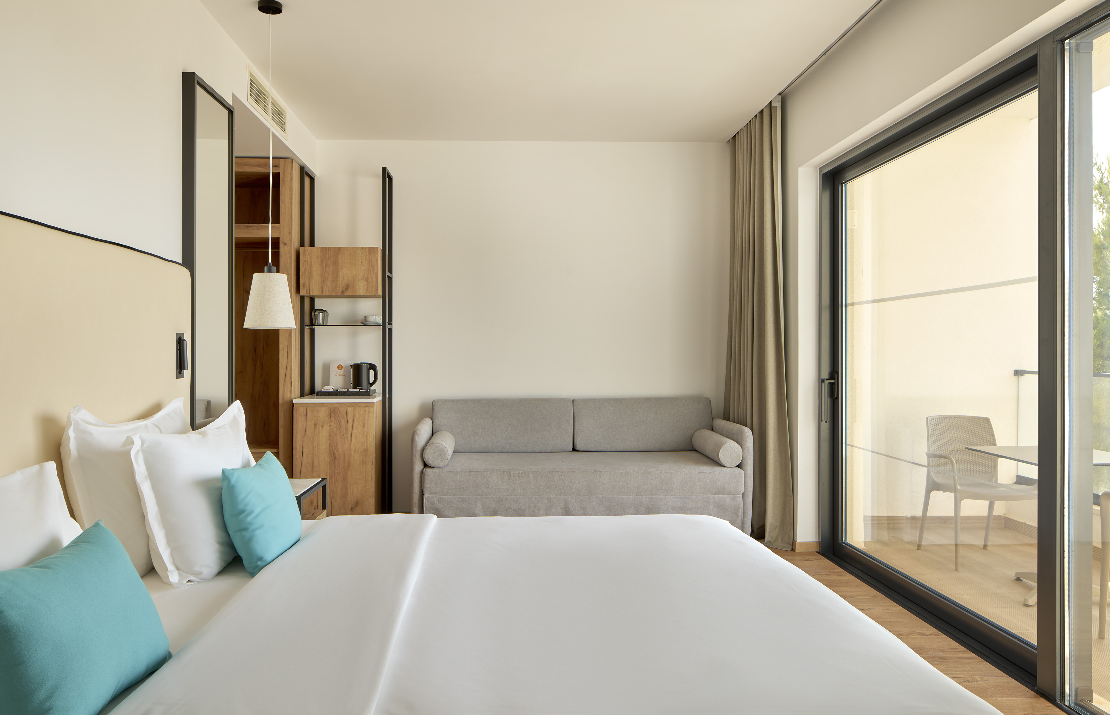 Bright modern hotel bedroom with white bed, gray sofa bed, and balcony view.