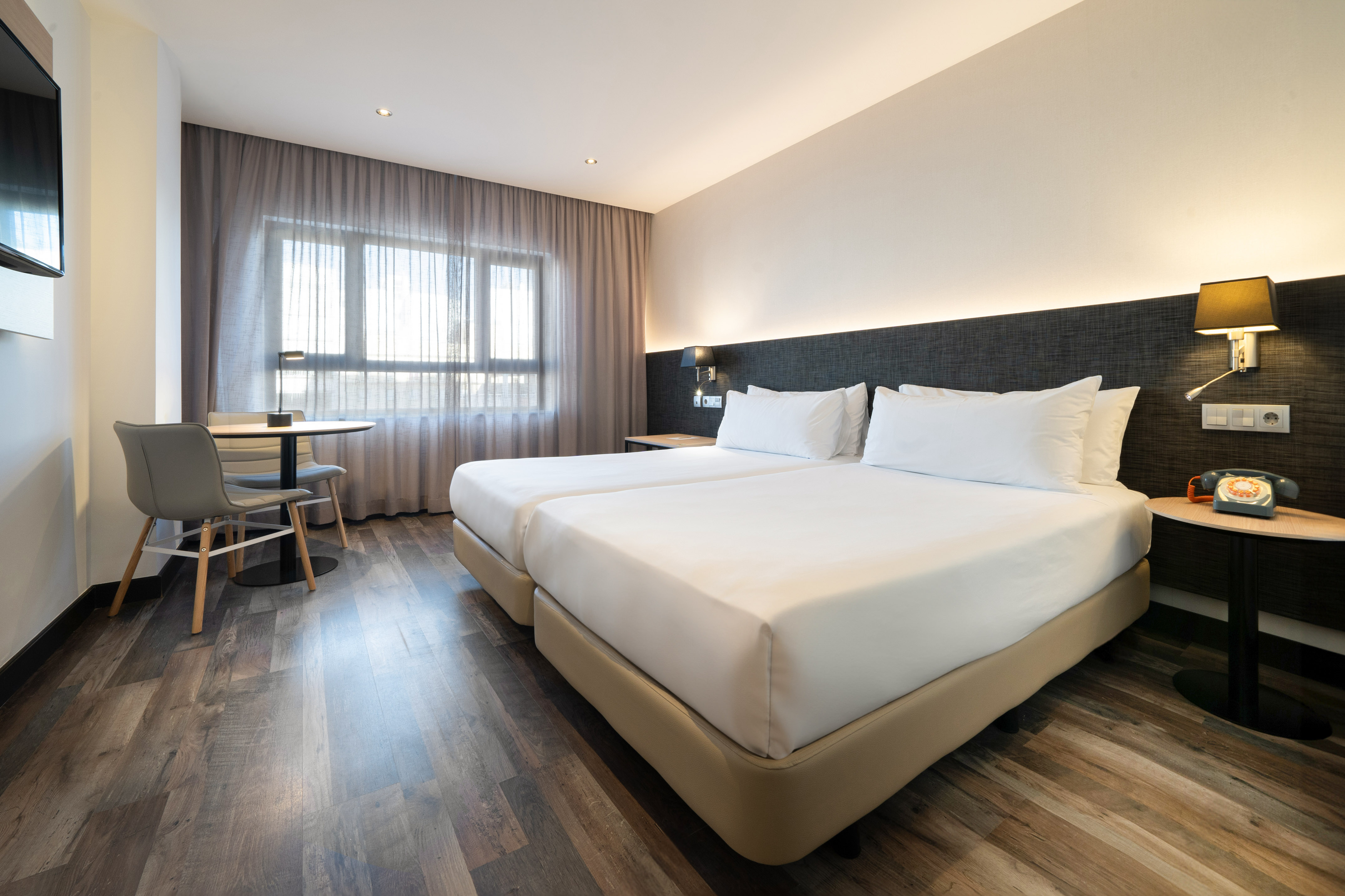 Modern hotel room with twin beds, small table, chairs, and wood-look flooring.