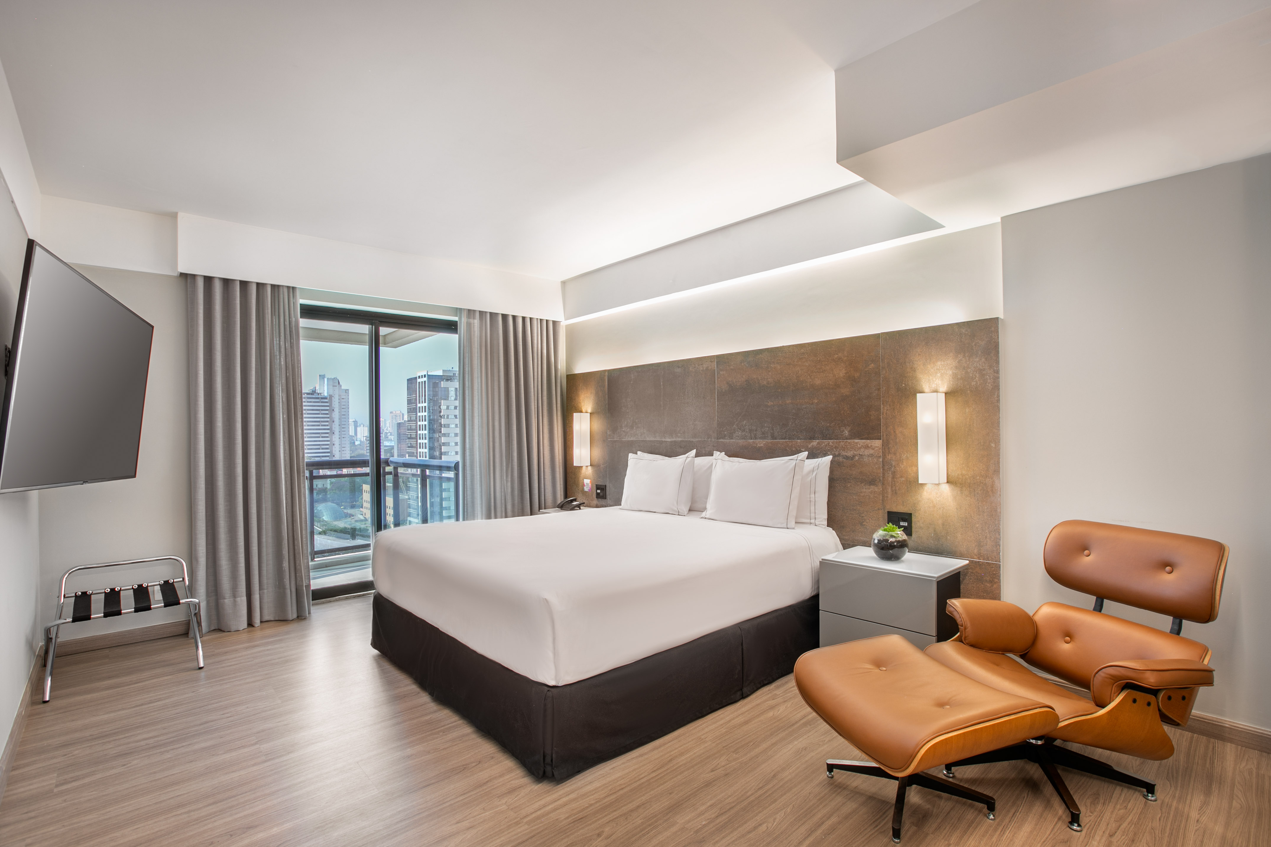 Modern hotel room with king bed, brown lounge chair, TV, and city view.