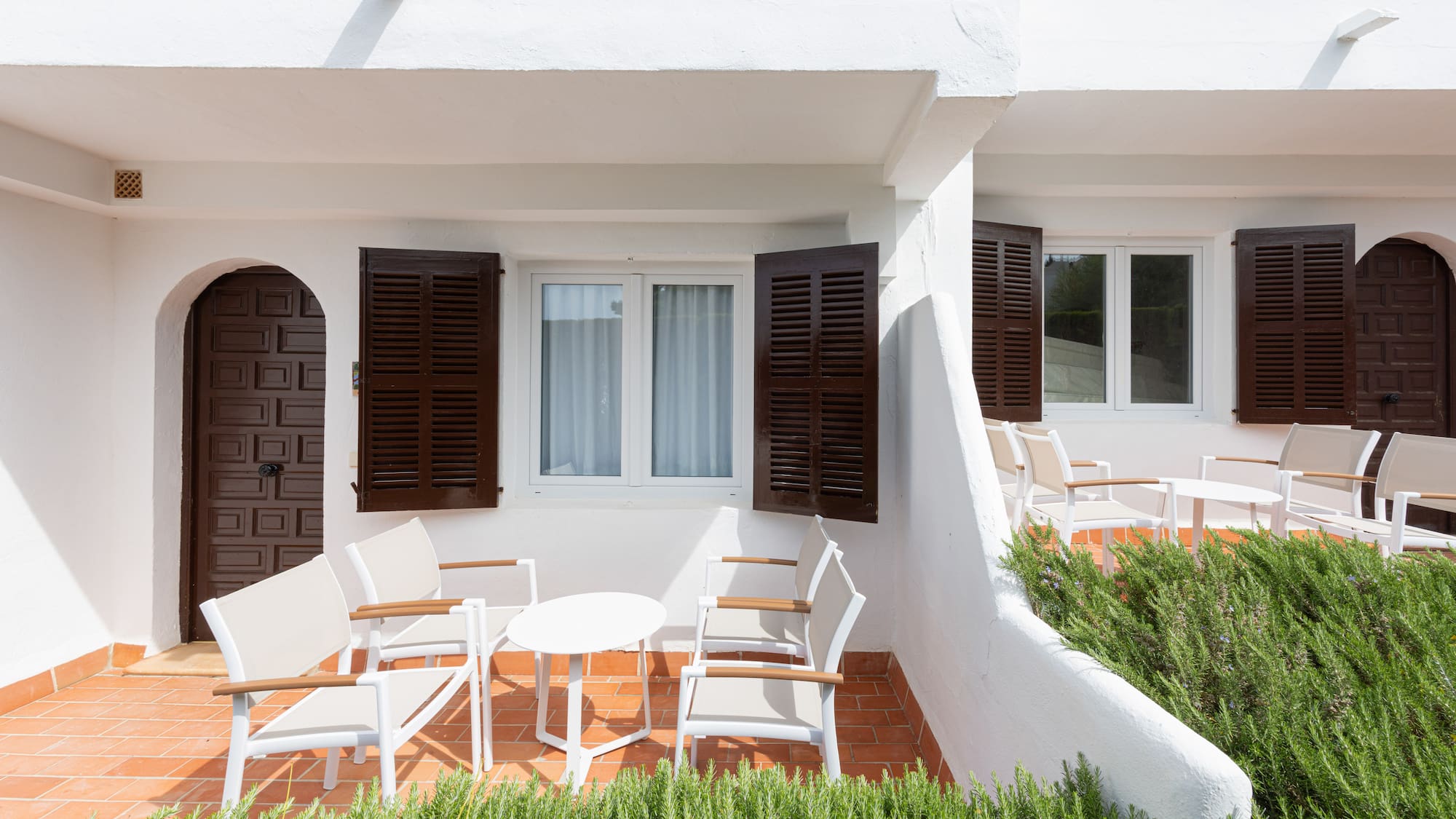 White hotel patio with terracotta floor, brown shutters, outdoor seating.