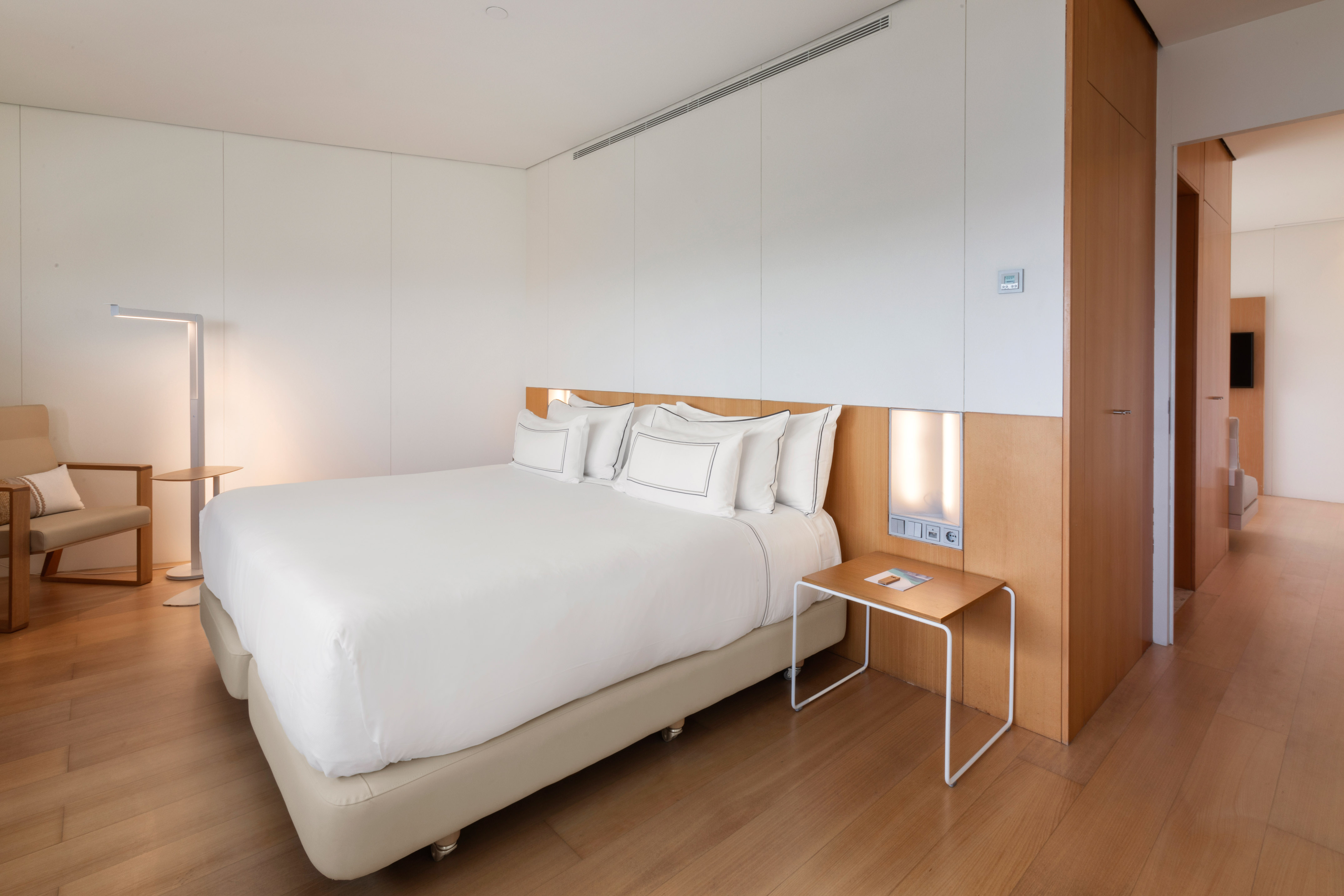 Clean, minimalist hotel room with white bed, wooden headboard, and light wooden floor.