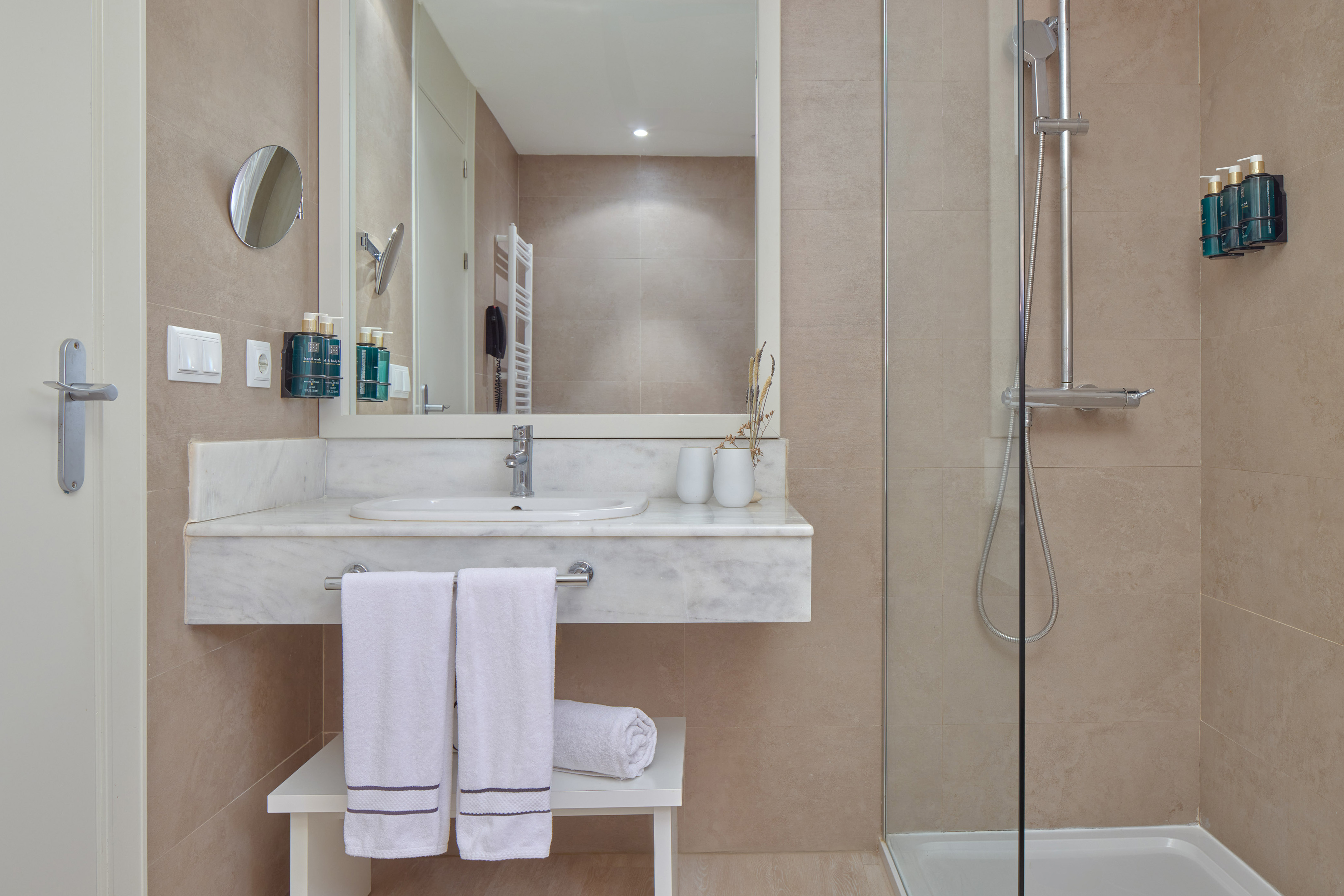 Modern hotel bathroom with marble vanity, white towels, glass shower, beige tiles.