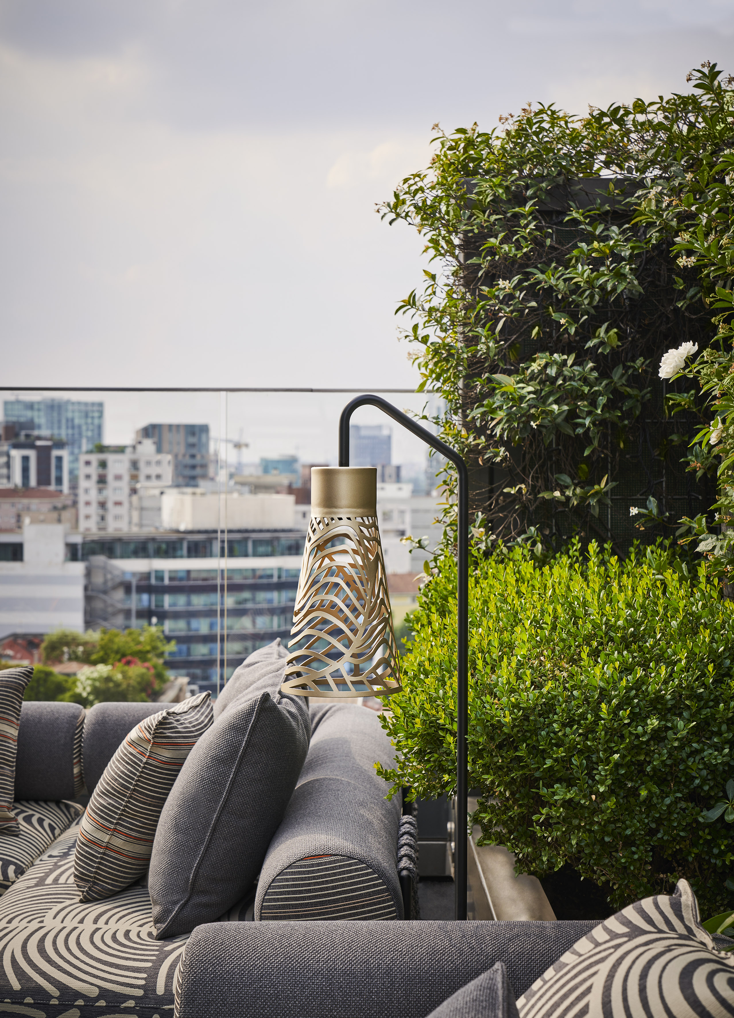 Outdoor terrace with modern sofa, woven lamp, green plants, and city skyline.