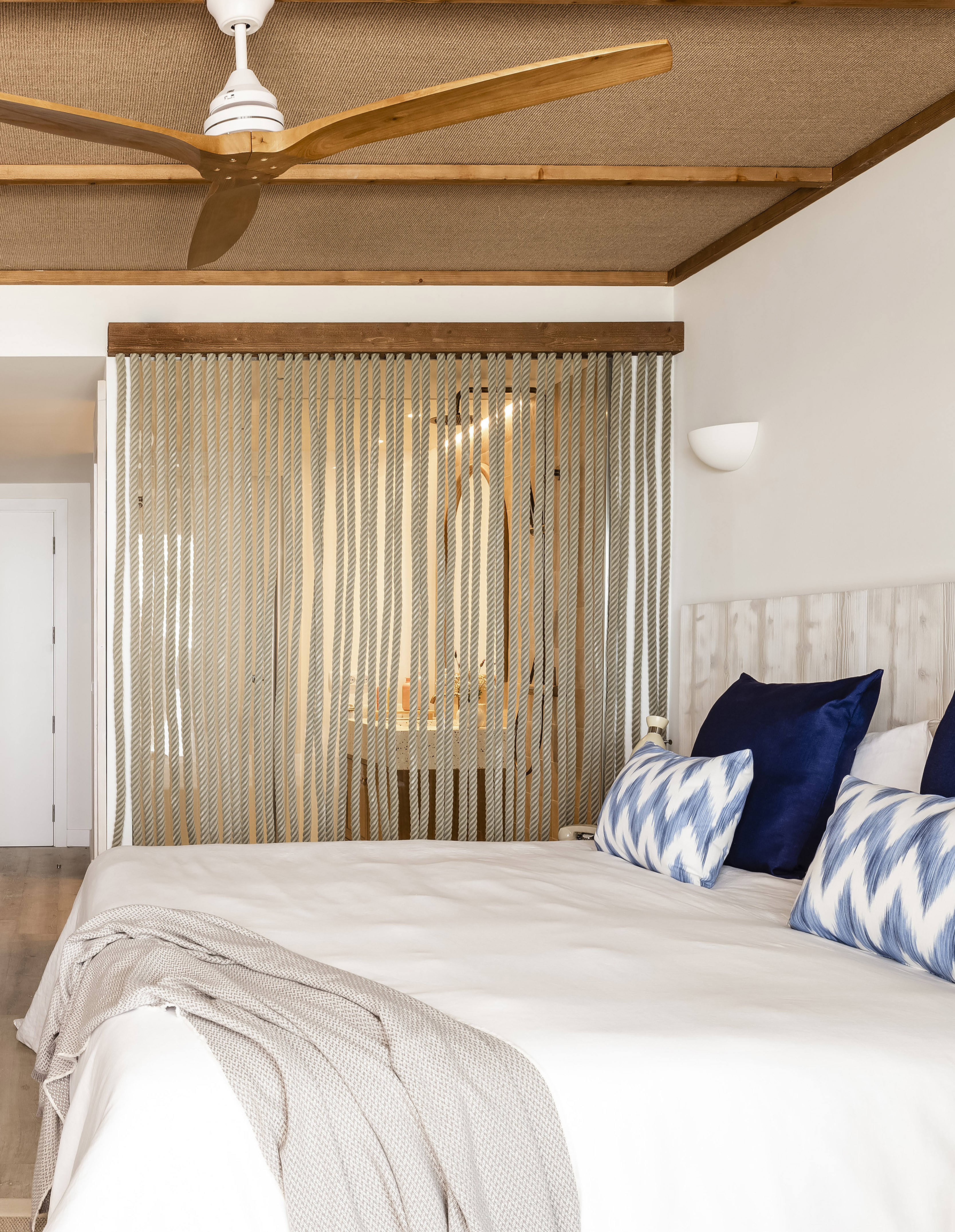 Tropical bedroom with white bed, blue pillows, wooden ceiling fan, rope screen.