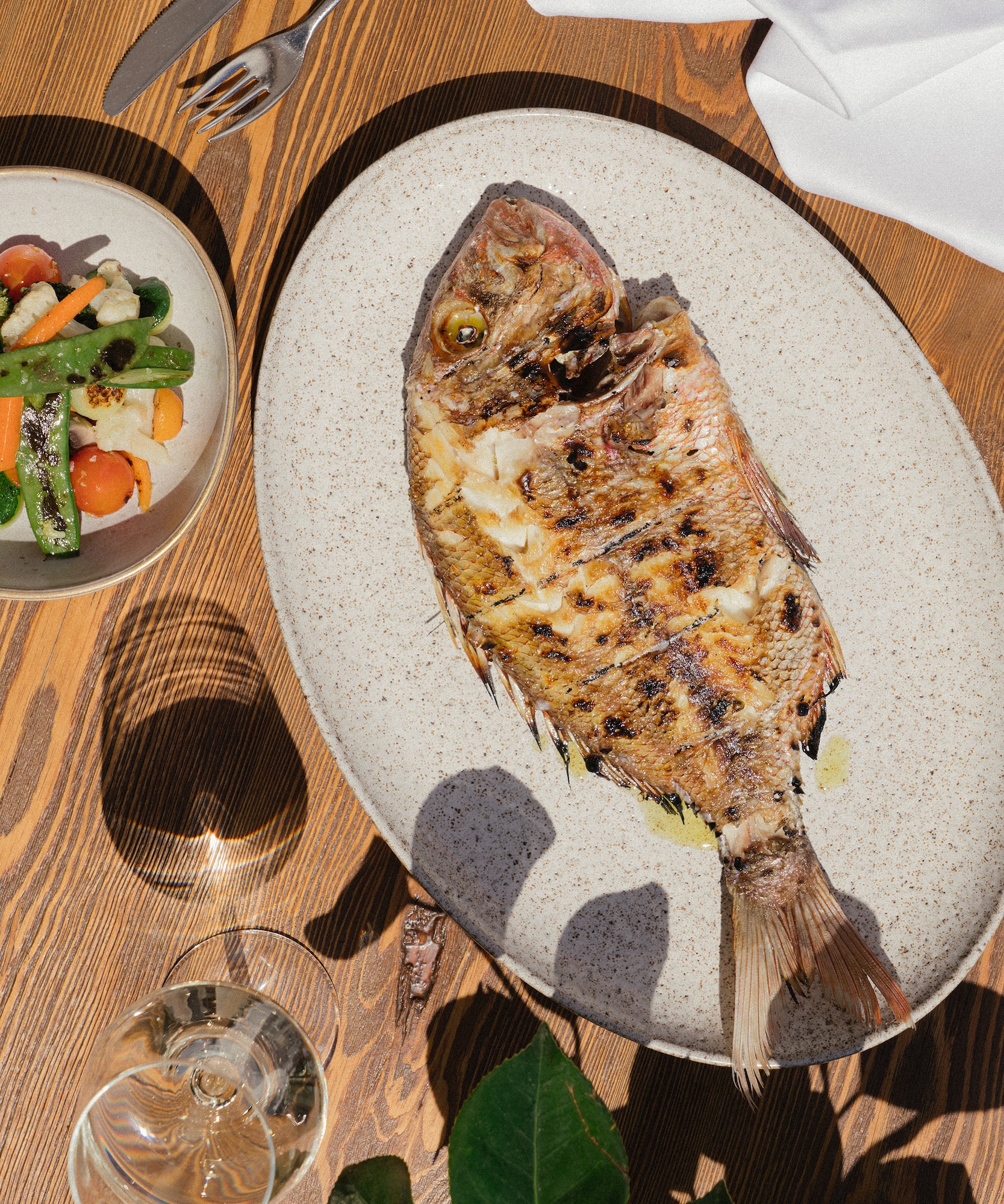 Pescado entero a la parrilla, verduras y copa de vino en mesa de madera.