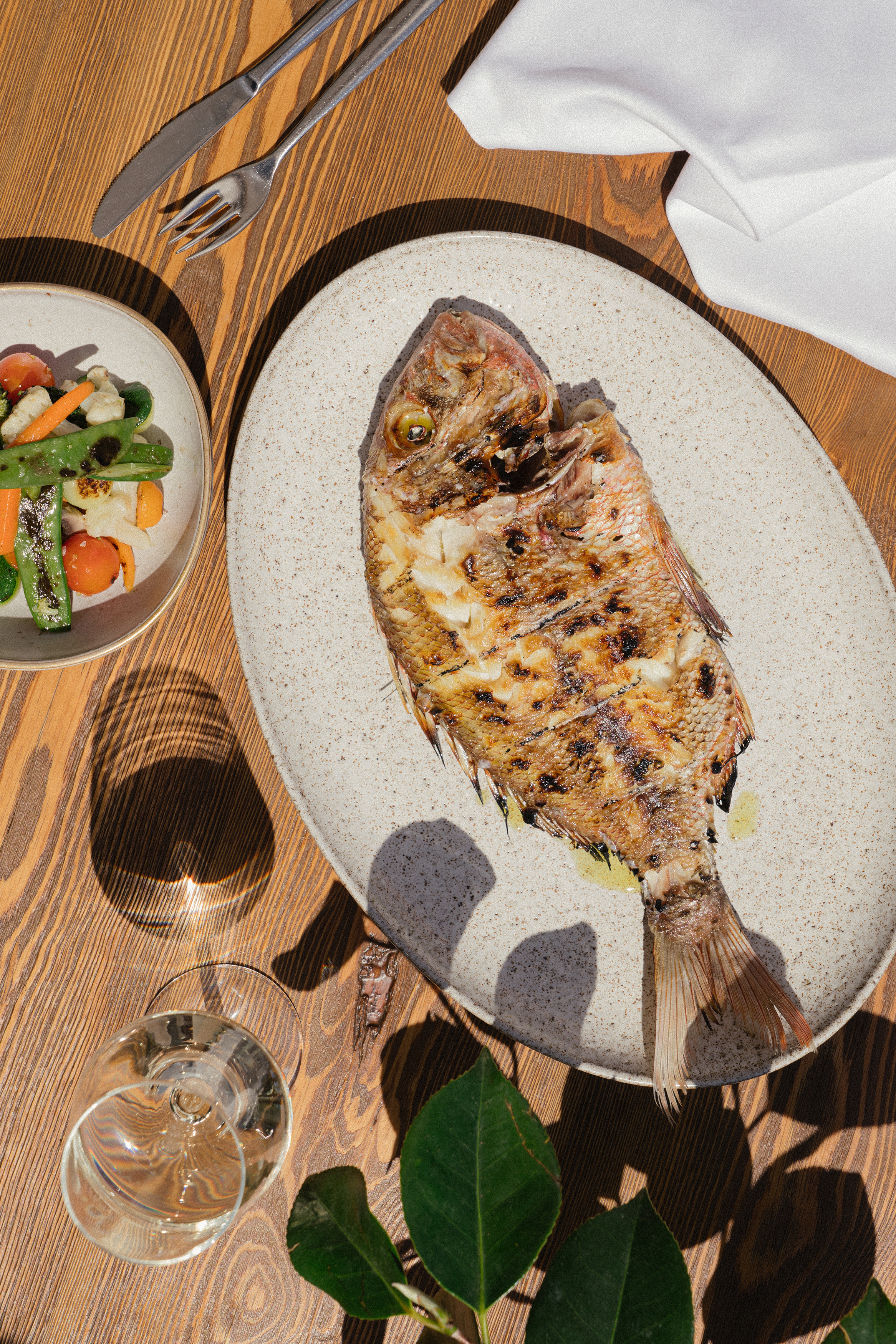 Pescado entero a la parrilla, verduras y copa de vino en mesa de madera.