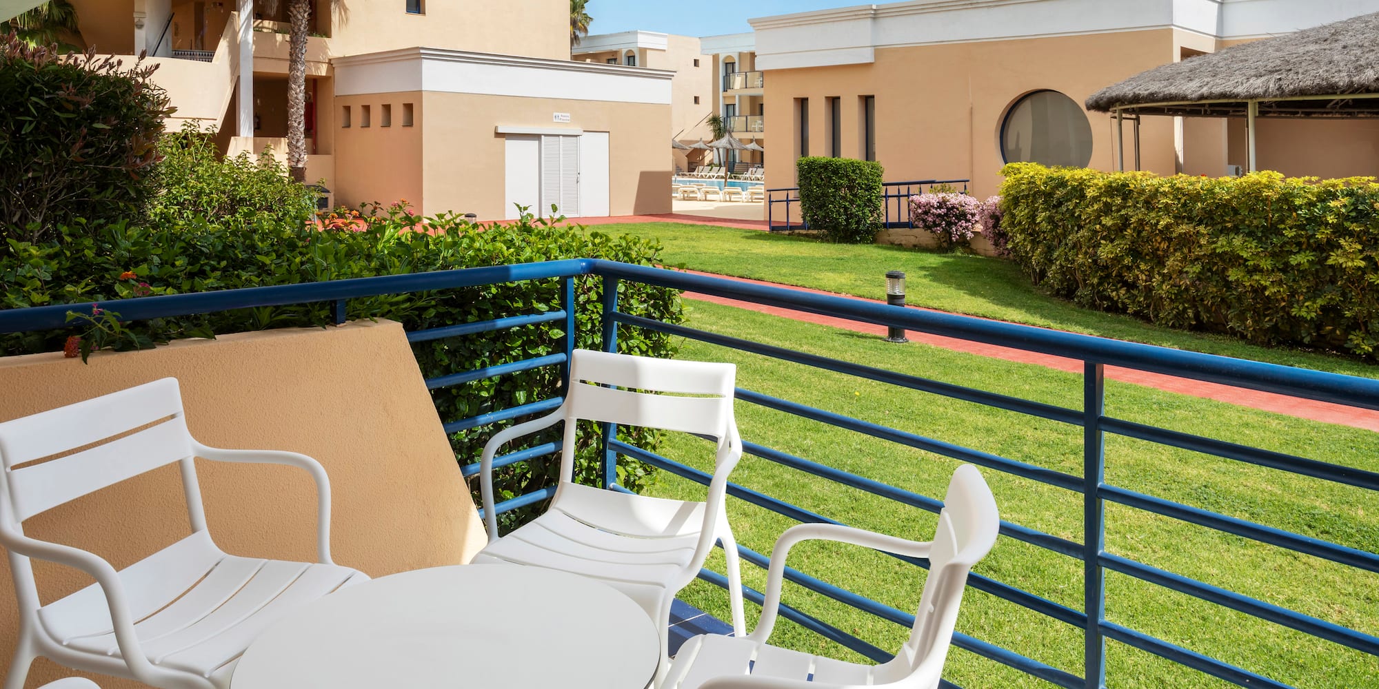 Resort balcony with white furniture, green lawn, buildings, and distant pool.
