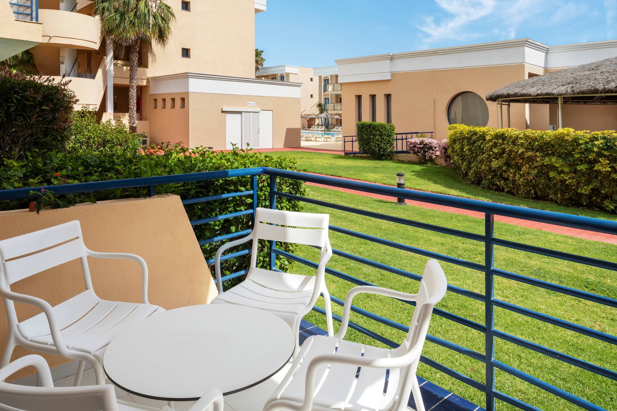Resort balcony with white furniture, green lawn, buildings, and distant pool.