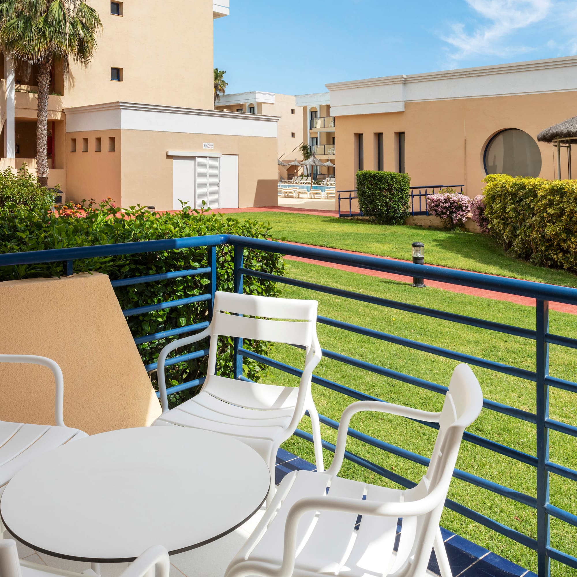 Resort balcony with white furniture, green lawn, buildings, and distant pool.