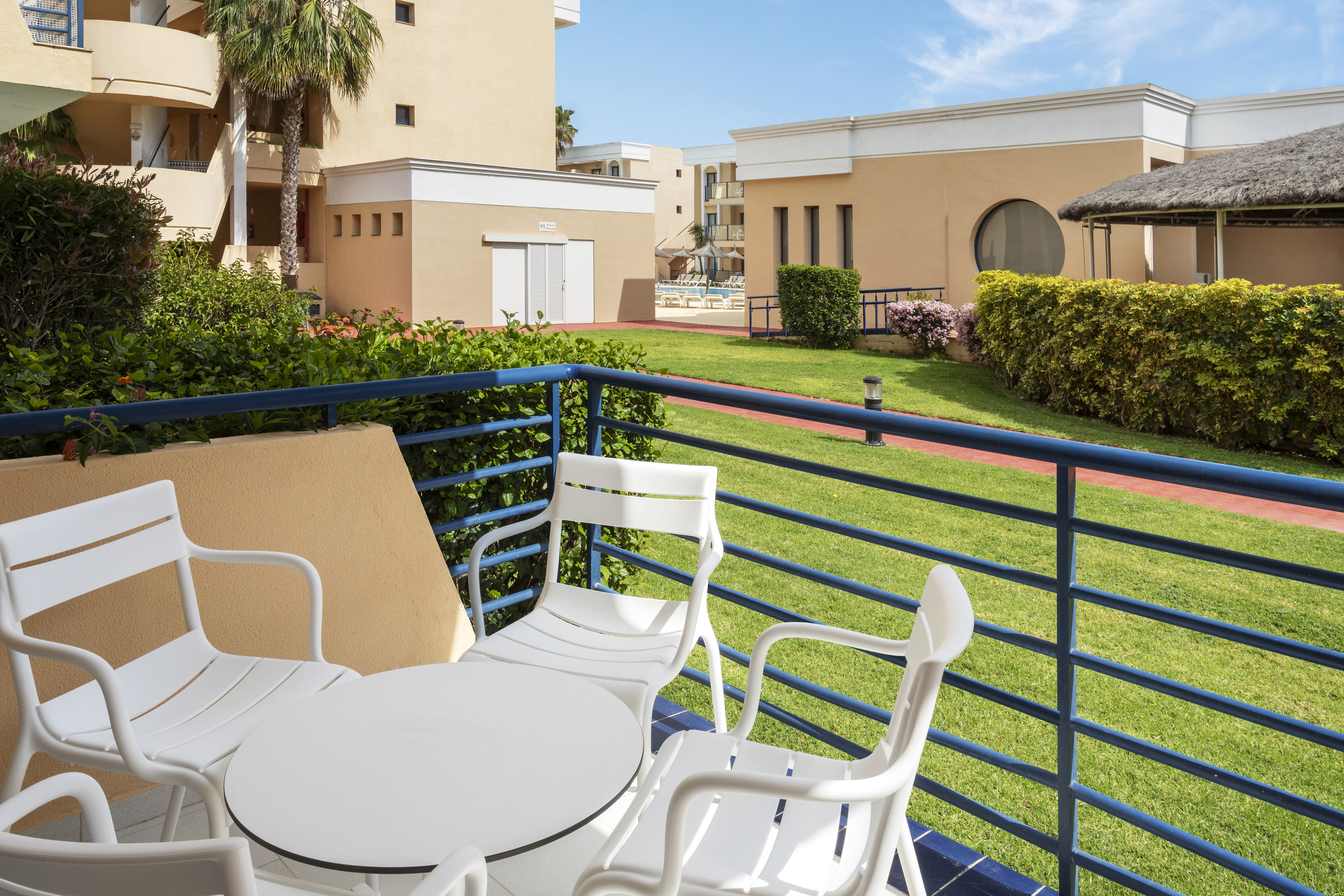 Resort balcony with white patio set, green garden, and distant pool.