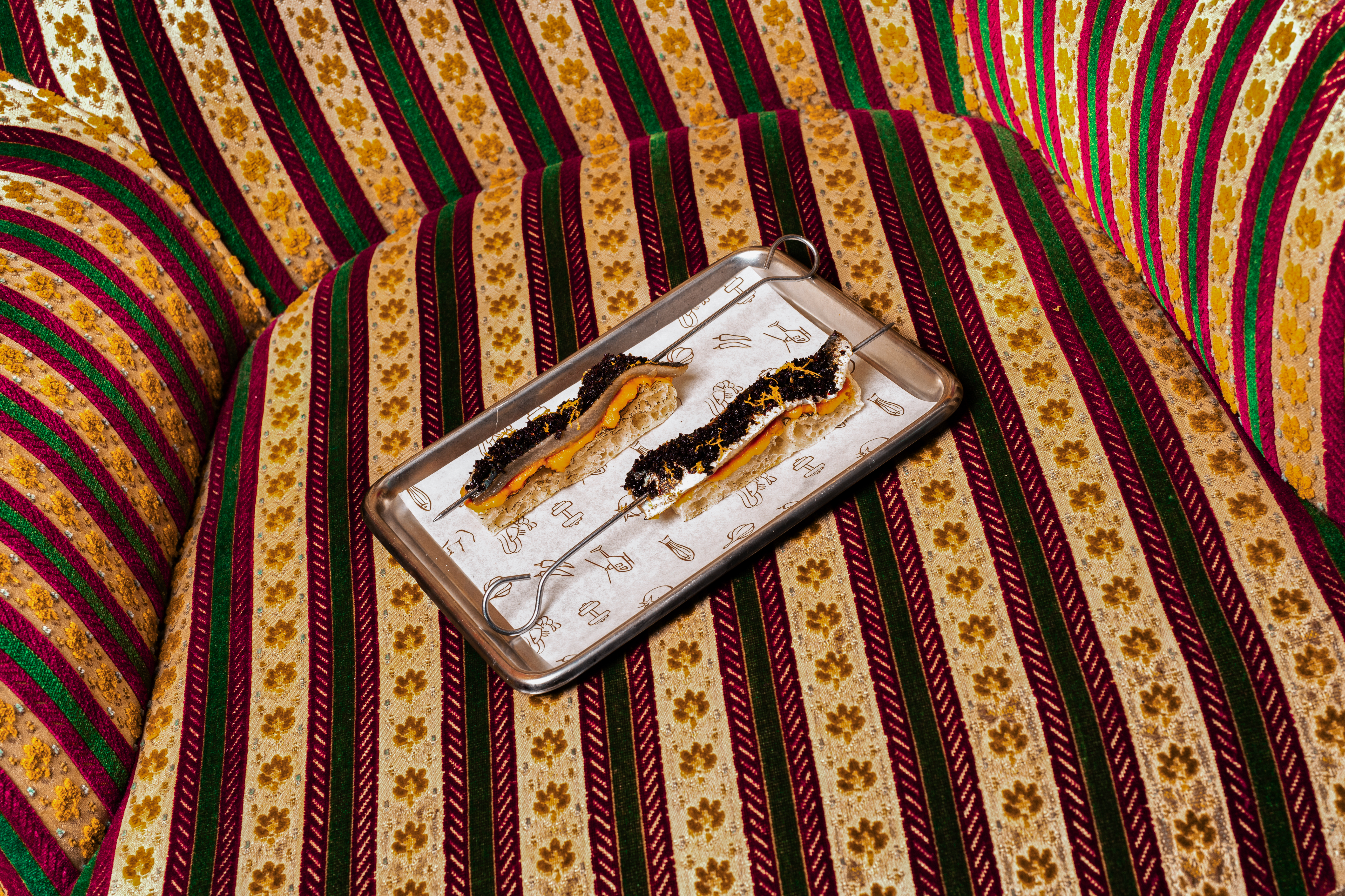 Canapés on skewers with dark sprinkles and orange zest, on a patterned sofa.