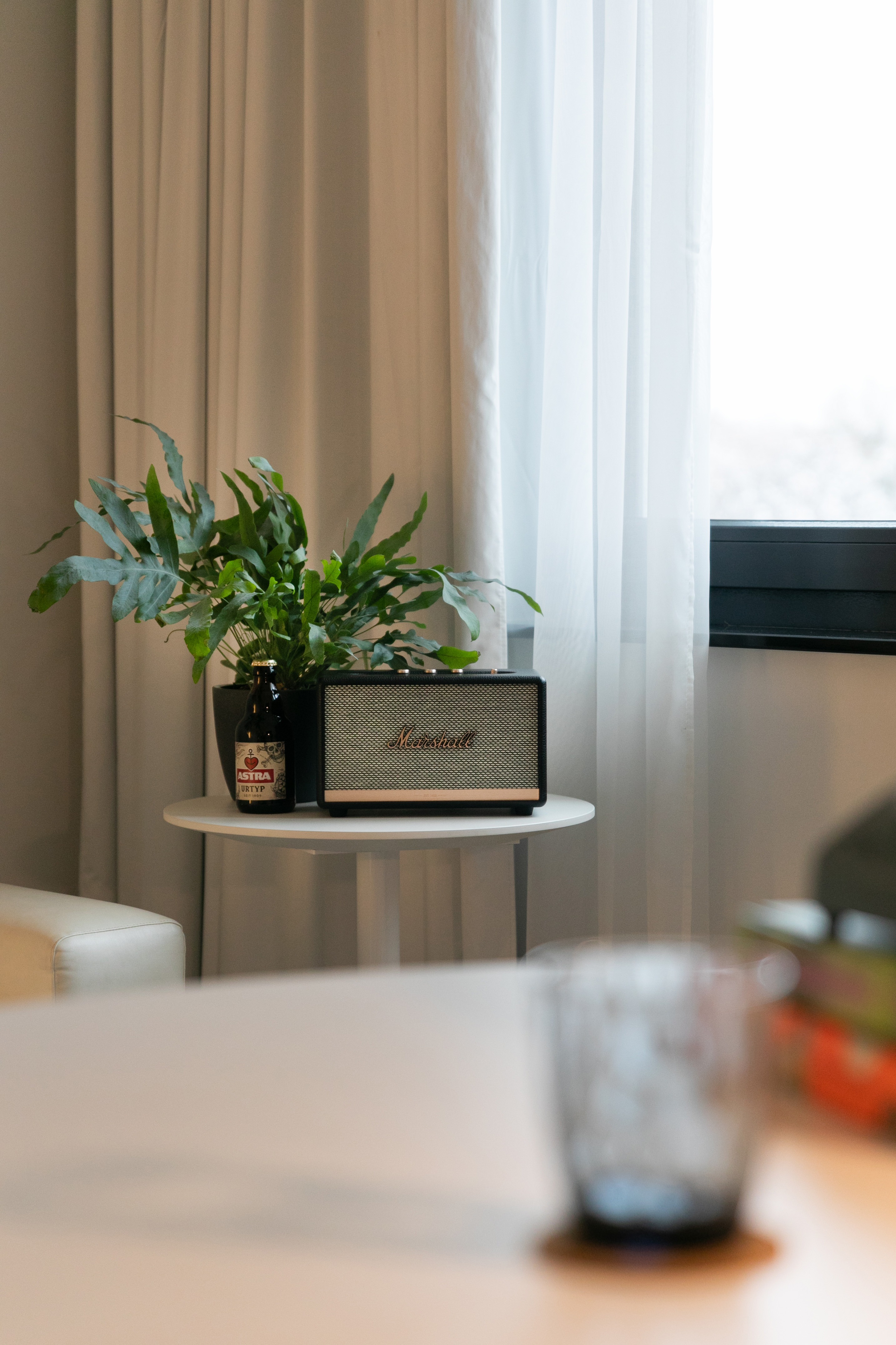 Marshall speaker, Astra beer, and plant on a white table next to sheer curtains.