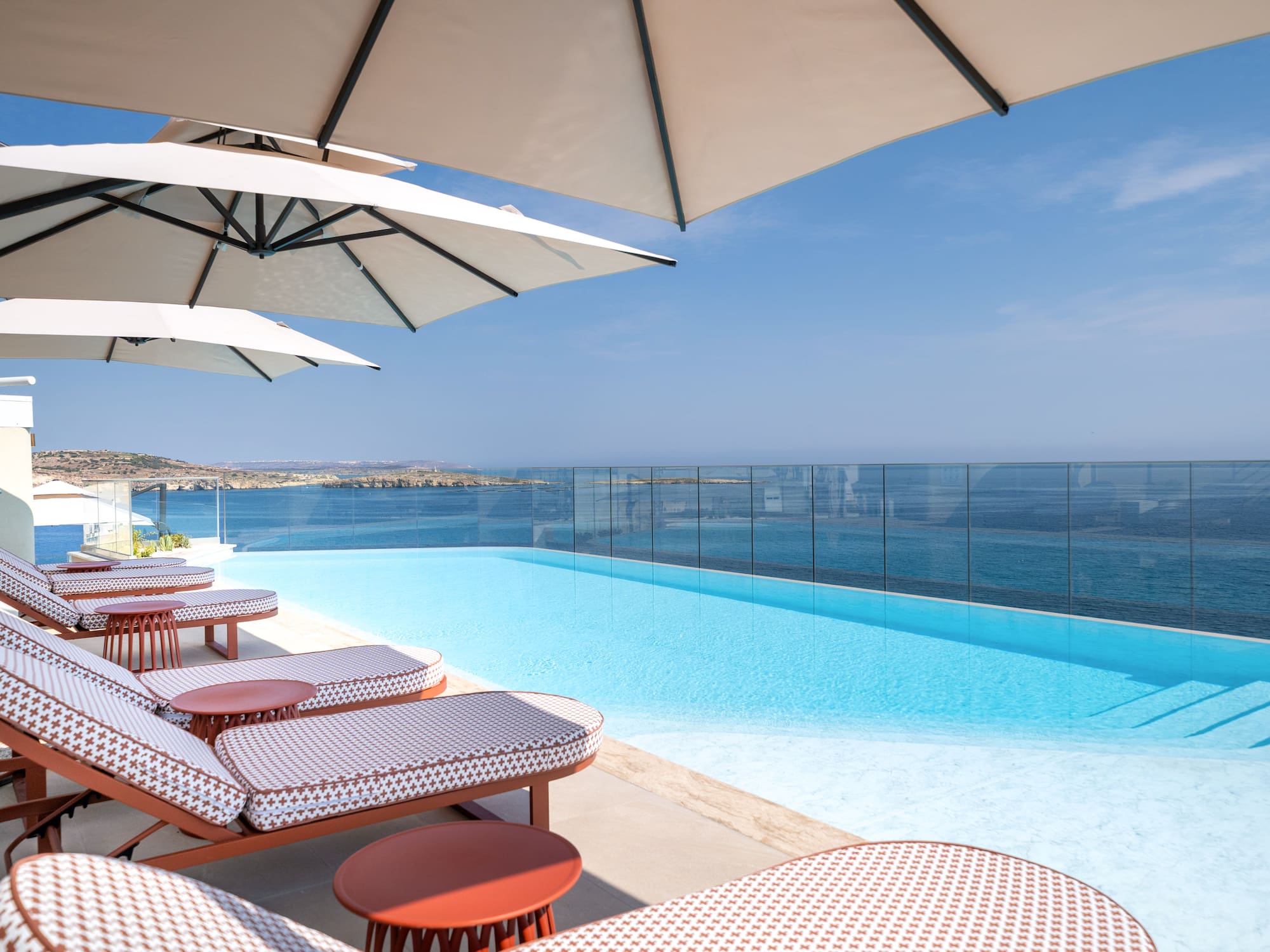 Infinity pool with ocean view, lounge chairs, and sun umbrellas on a sunny terrace.