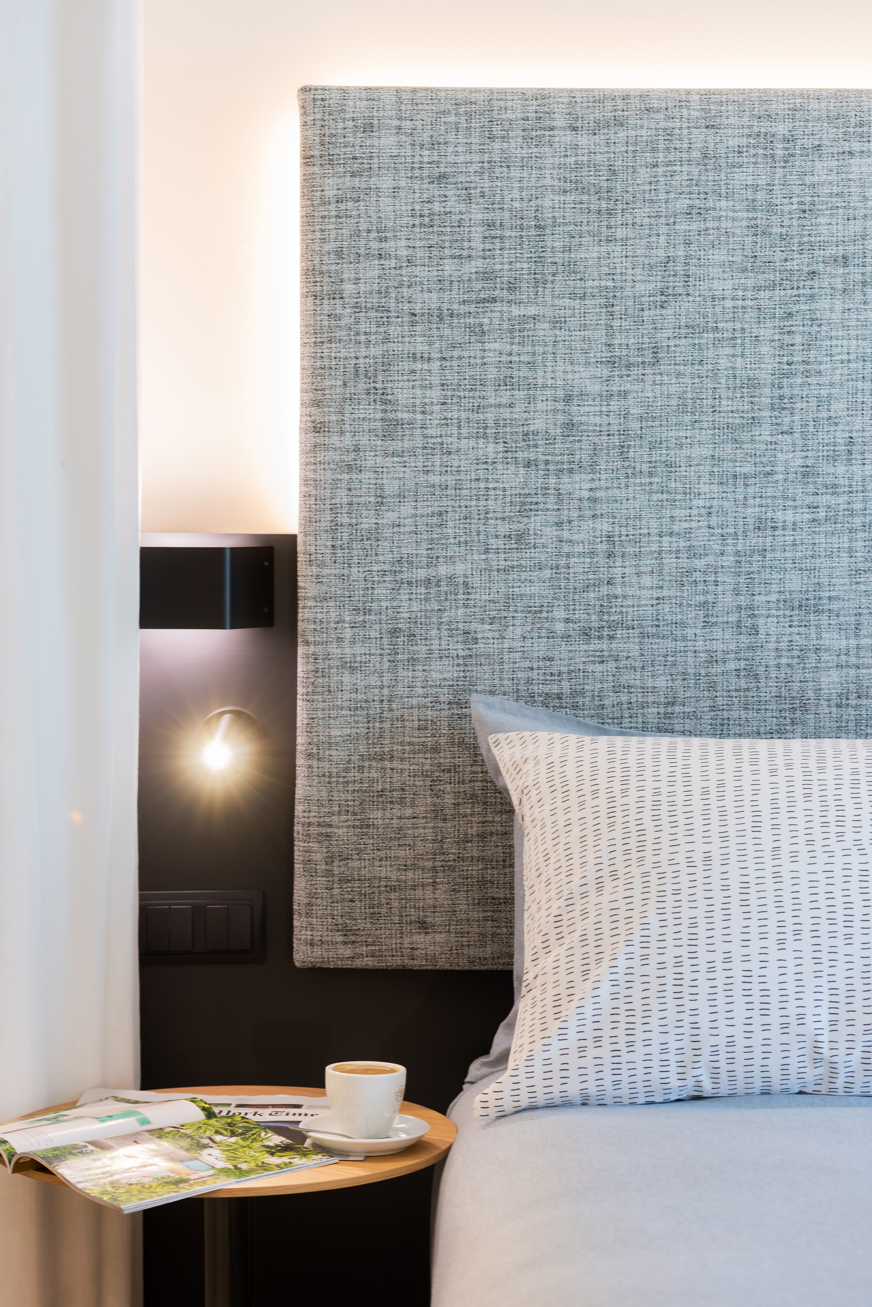 Modern bedroom with grey headboard, bedside lamp, coffee, and magazine.