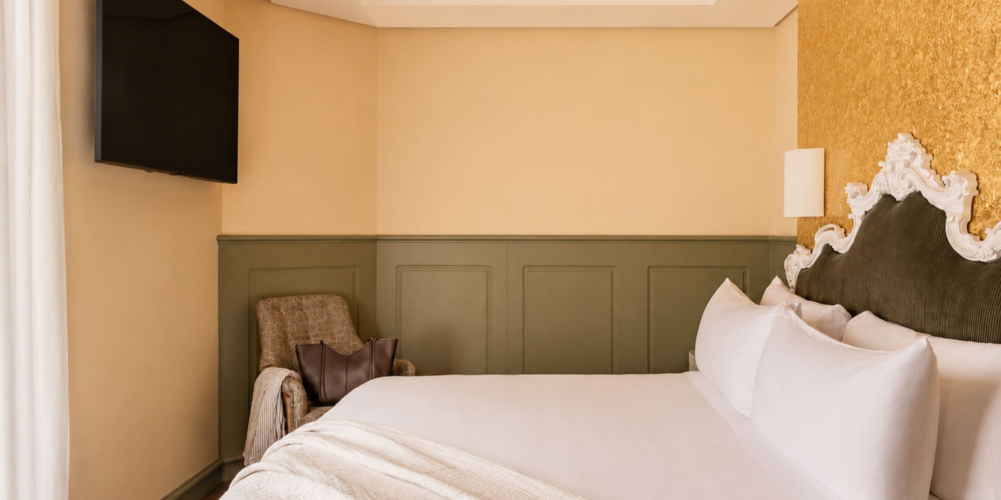 Elegant hotel bedroom with white bed, ornate headboard, gold accent wall, and TV.